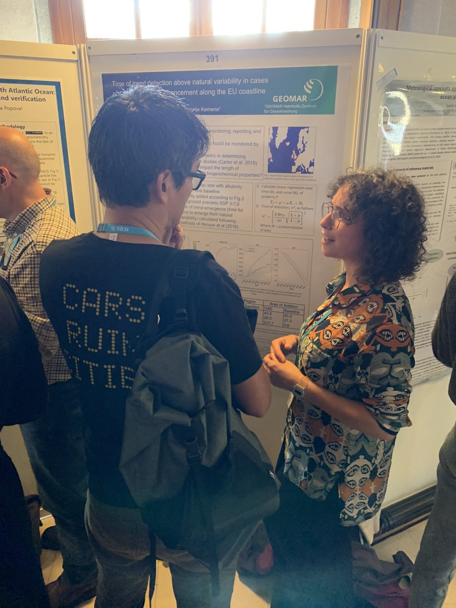 Two people are engaged in conversation in front of a research poster at a conference. One person is wearing a black T-shirt with yellow text that says Cars Ruin Cities, while the other has curly hair and is wearing a colorful patterned shirt. The poster discusses trends and variability in detectability of ocean alkalinity enhancement along the EU coastline