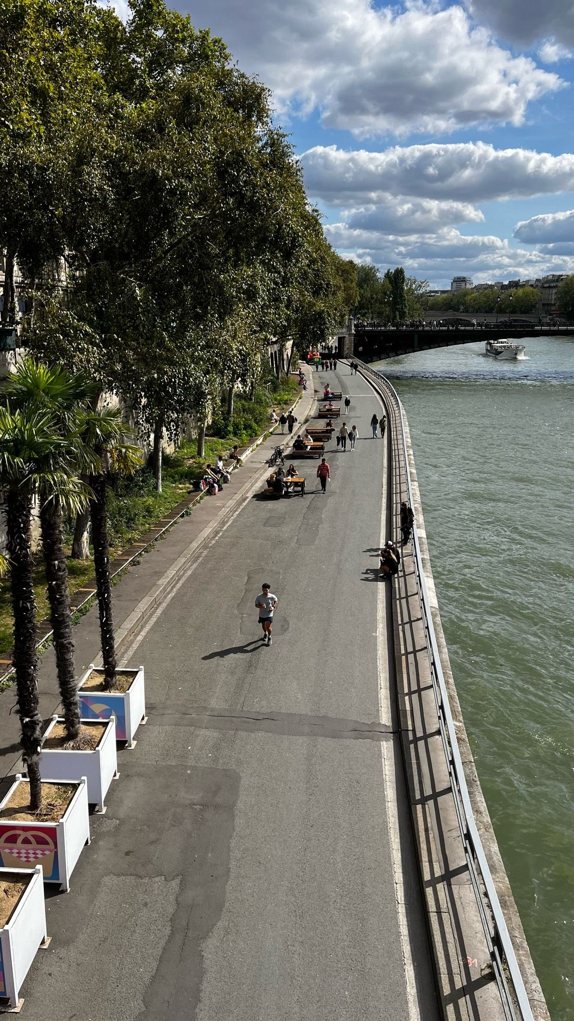 A riverside pathway lined with palm trees and planters, showing people walking, sitting, and cycling. A boat is visible on the water, with a bridge in the background under a partly cloudy sky.