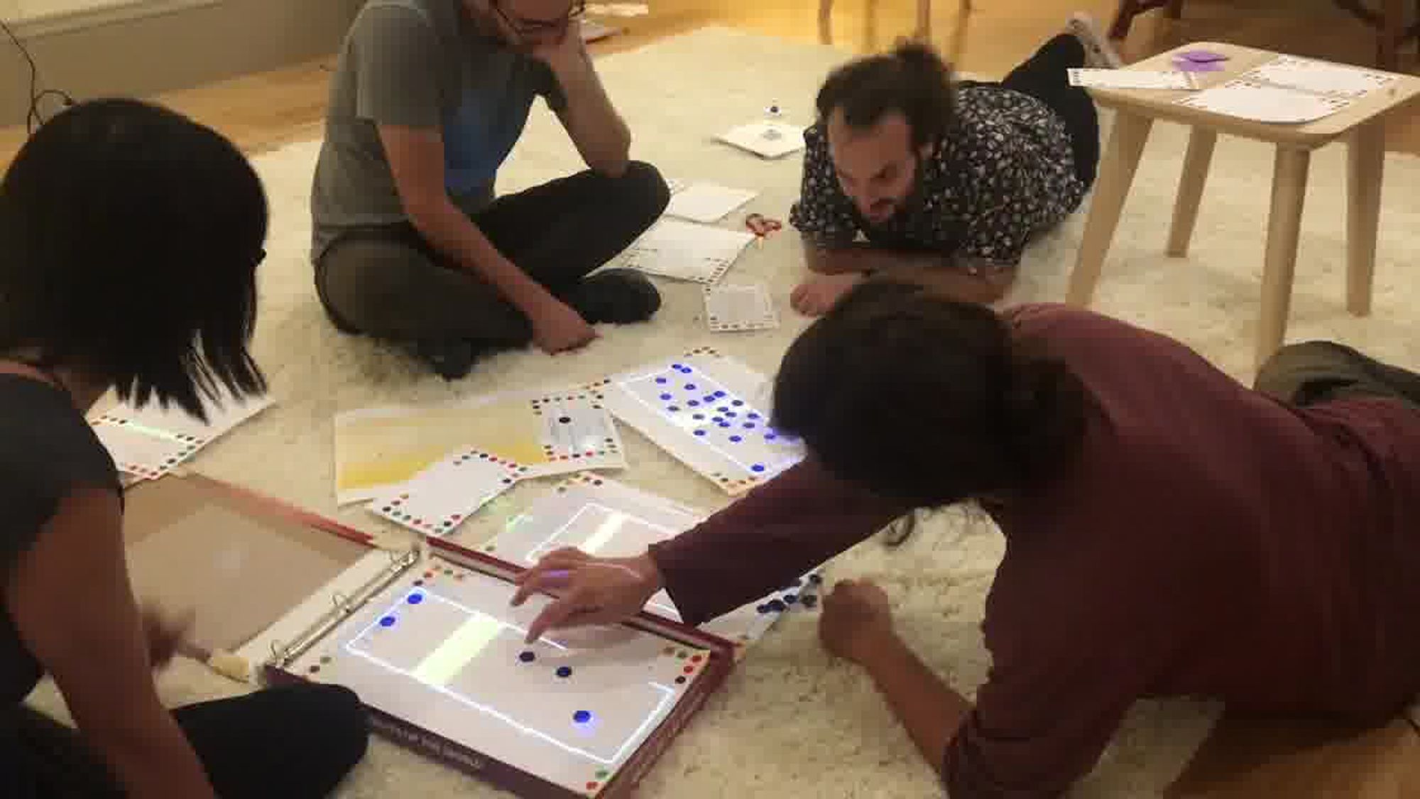 A photograph of four people sitting or lying on the floor, gathered around a set of Dynamicland programming cards and projected images, working together on a common project.