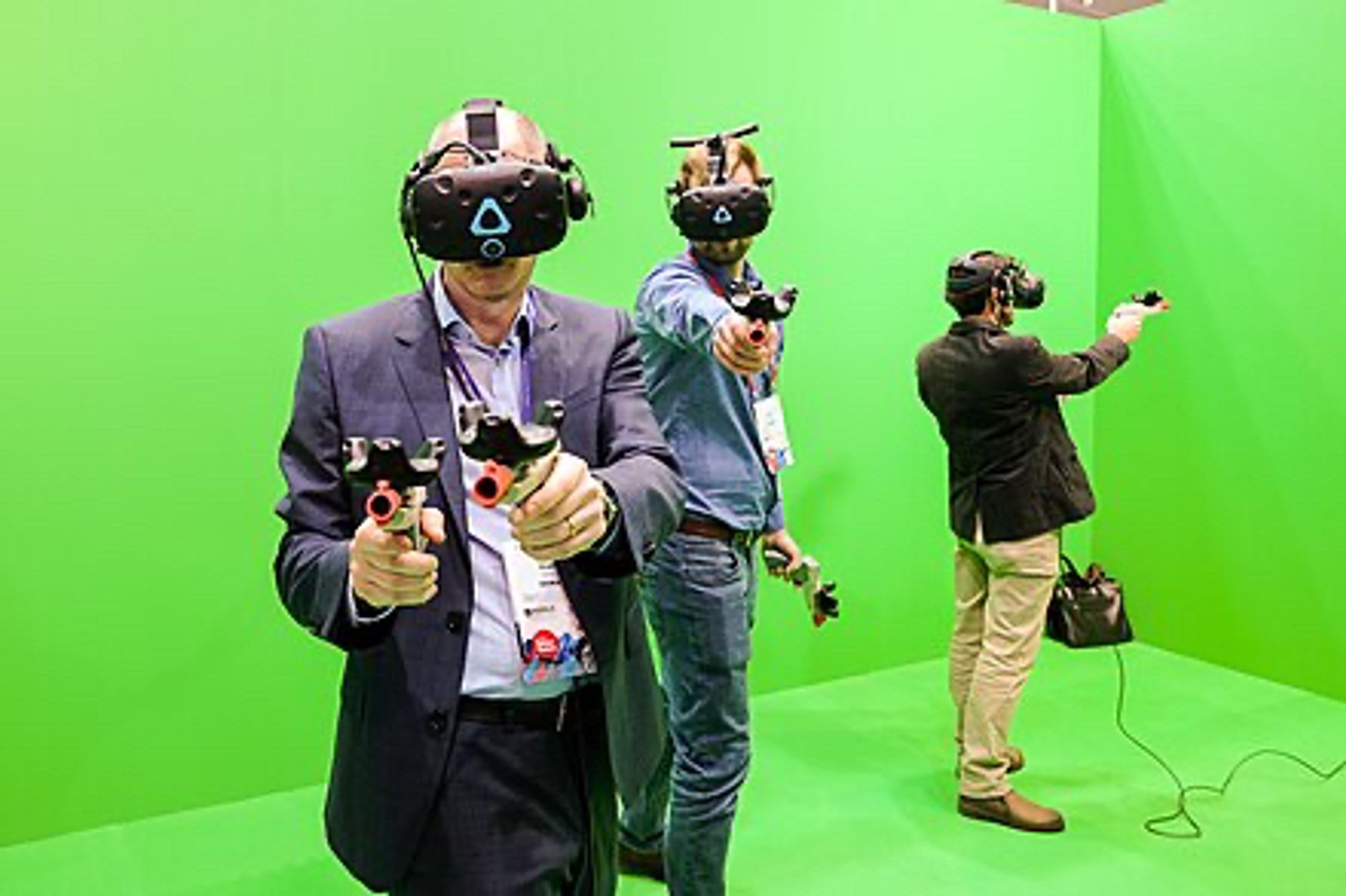 A picture of three men using virtual reality headsets that cover their faces. They are holding gun-like devices that they can point to various places in a room with green-screen walls.