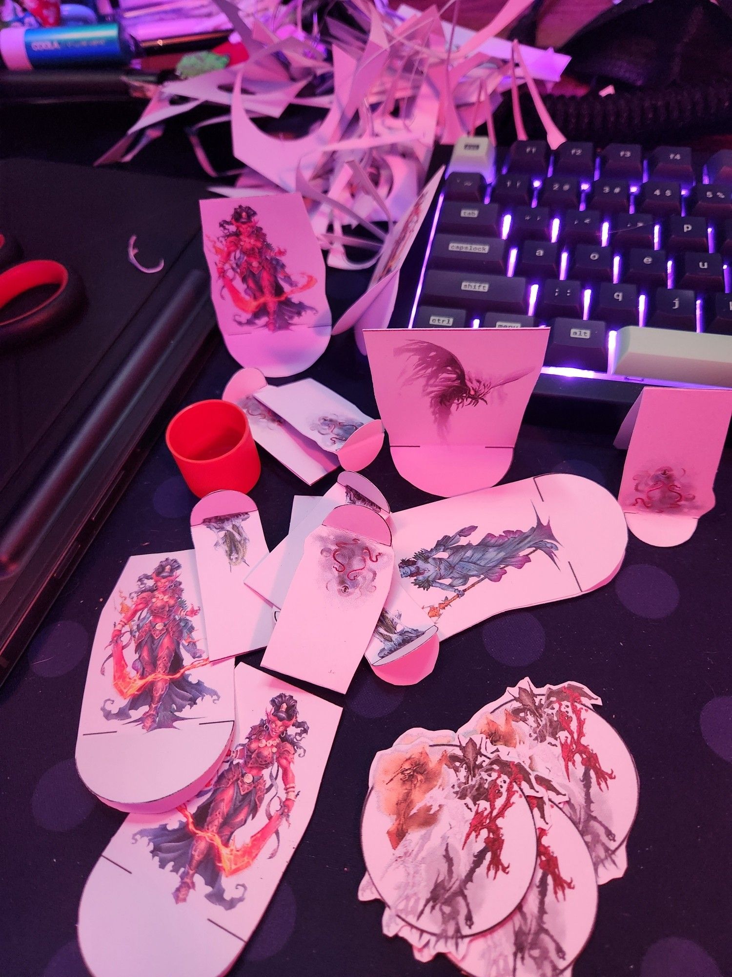 photegraph of jess' desk, primarily of printed off tokens and paper miniatures mid-asnebbly featuring artwork of multiple different elementals and genies. Behind the paper miniatures is jess' keyboard and a mound of paper cutting scraps.