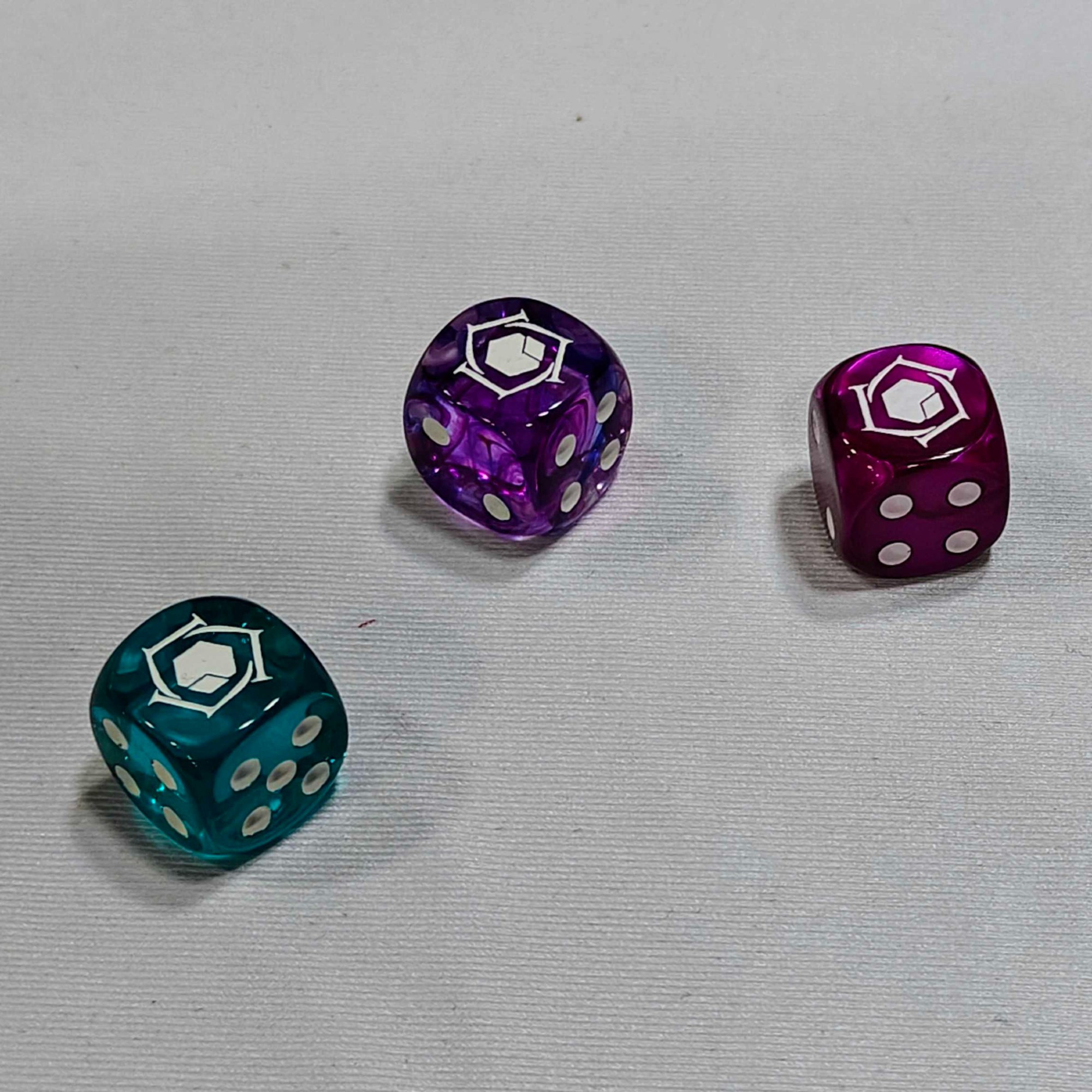 3 six-sided die with the Lore Link logo. One is a greenish-blue, one is purple, and one is a marbled reddish-purple. Merlot, perhaps?