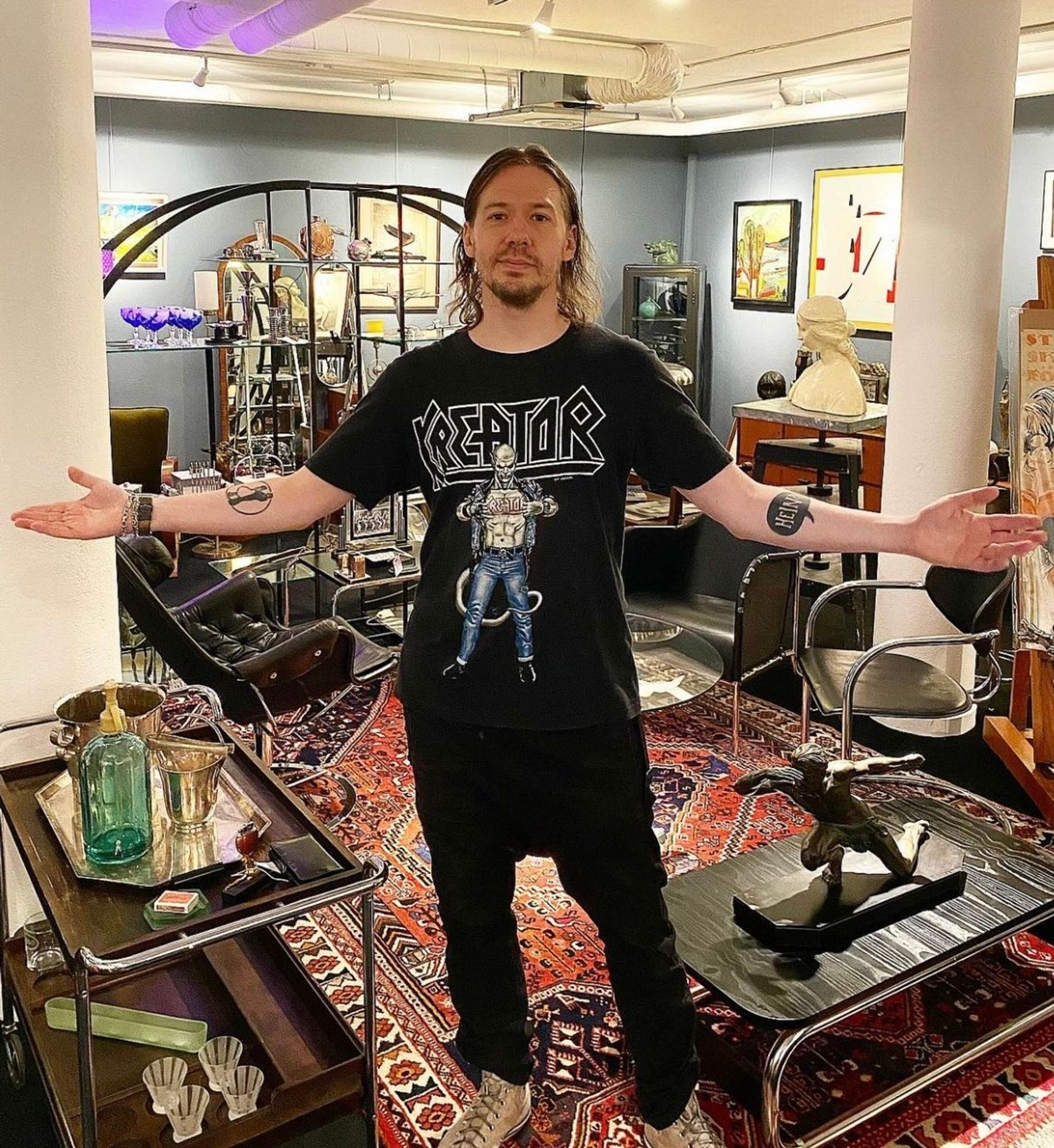 Tobias Forge, vocalista e líder da banda sueca Ghost, em uma foto em uma sala com artefatos variados. Ele está no meio da foto usando uma blusa da banda Kreator, calça preta, tênis claro e está com os braços abertos como se estivesse recebendo alguém.