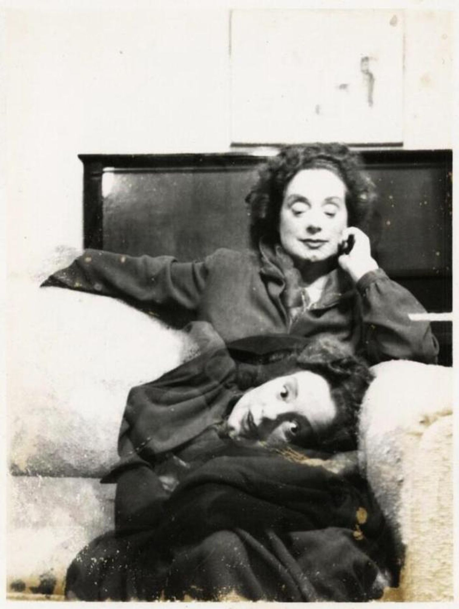 Black and white photography of a smiling Elsa Lanchester with a head dummy of hers on her lap, while sitting on a couch