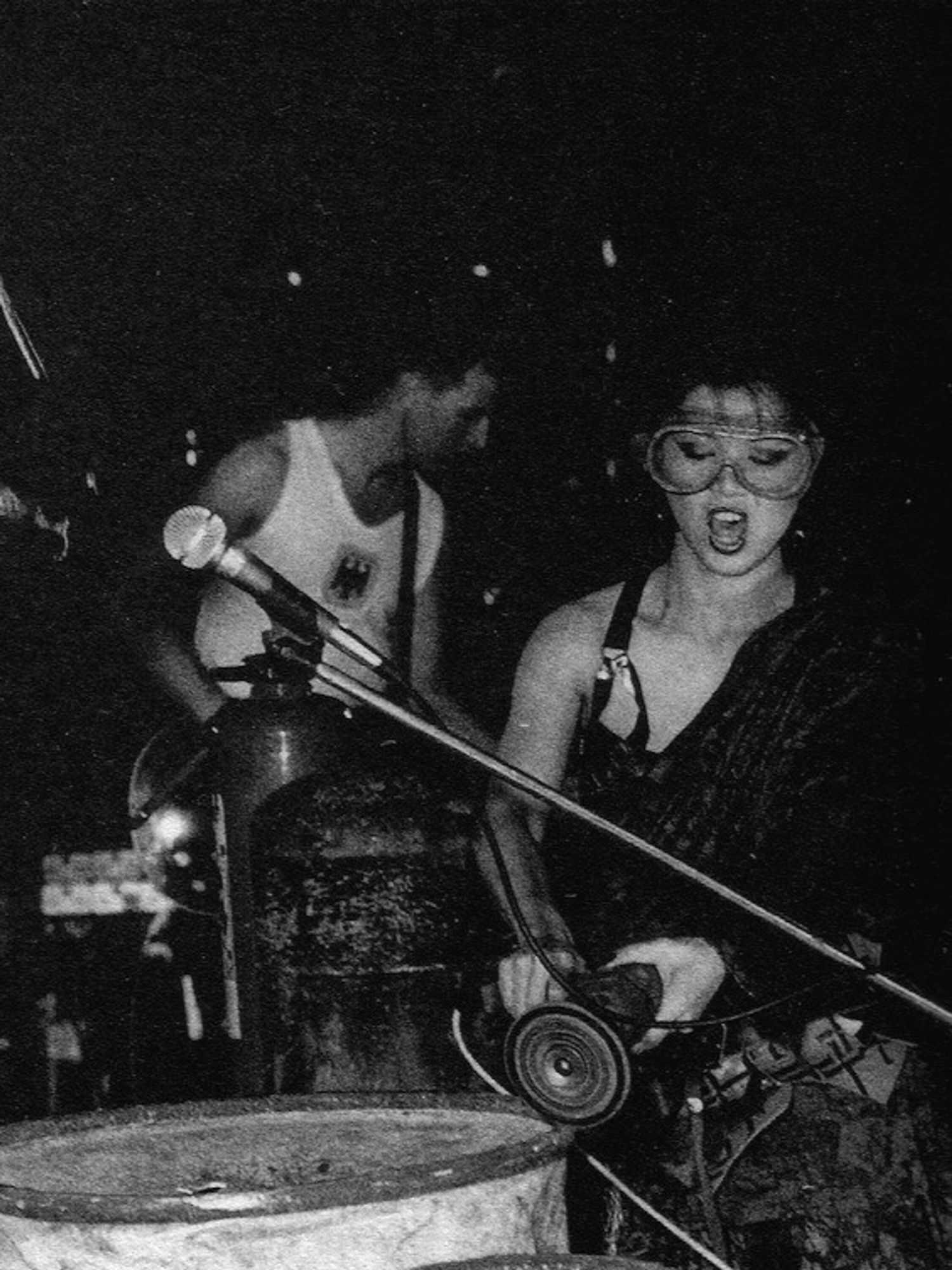 Black and white photo of a caucasian man in a German Bundeswehr undershirt playing what assumingly must be a bass, while an Asian woman wearing protective goggles is working an oil drum with a grinding machine, a microphone juts in the foreground