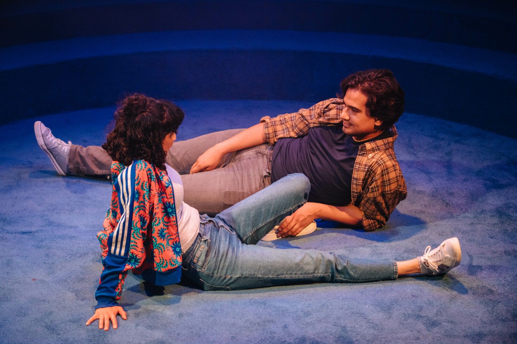 Mariam Haque as Neelam and Nathaniel Curtis as Zaid sit casually chatting on a blue carpet. The Real Ones at the Bush Theatre's Holloway, 2024.