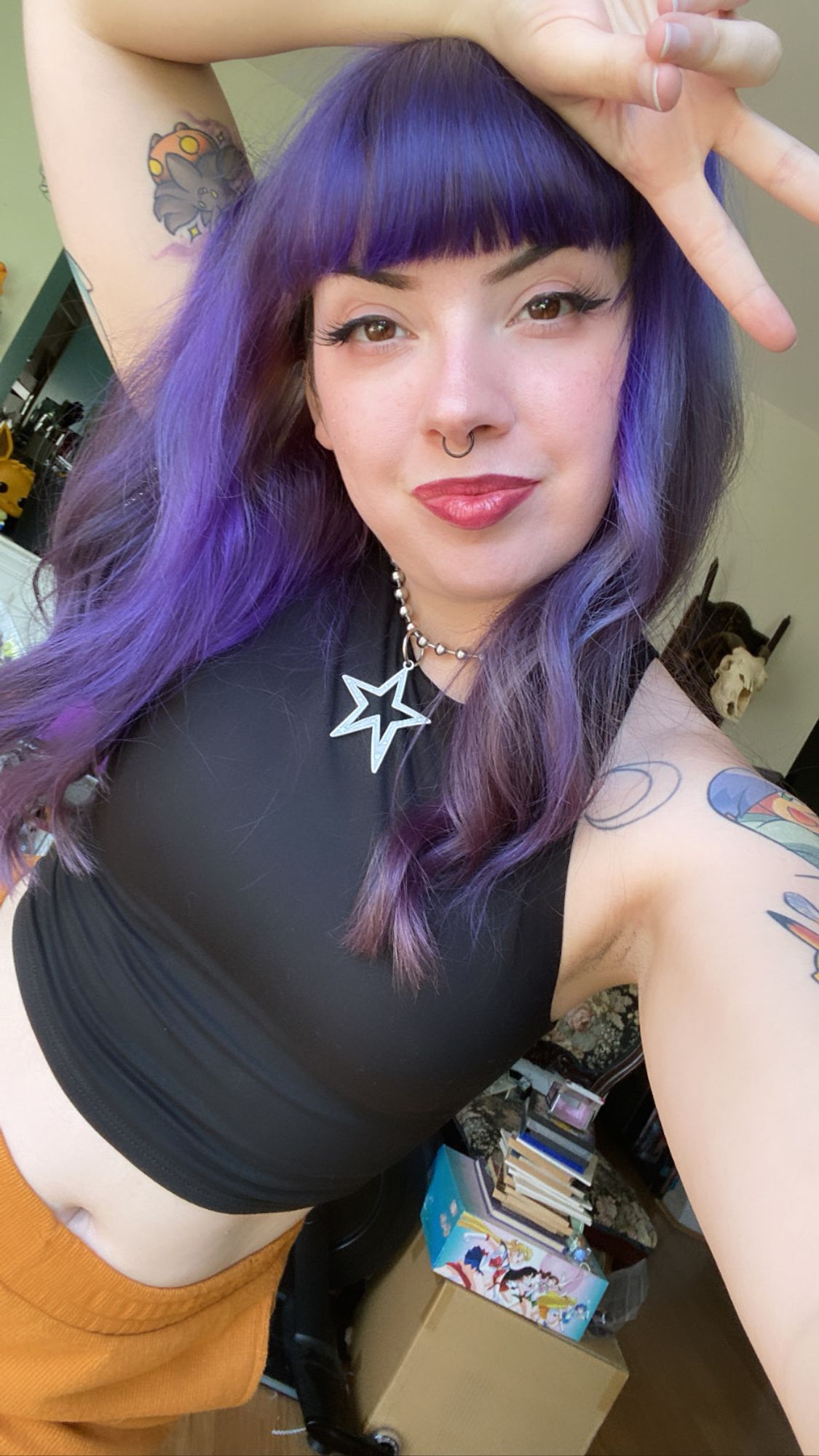 purple haired girl in a black tank top poses in her living room, throwing up a peace sign