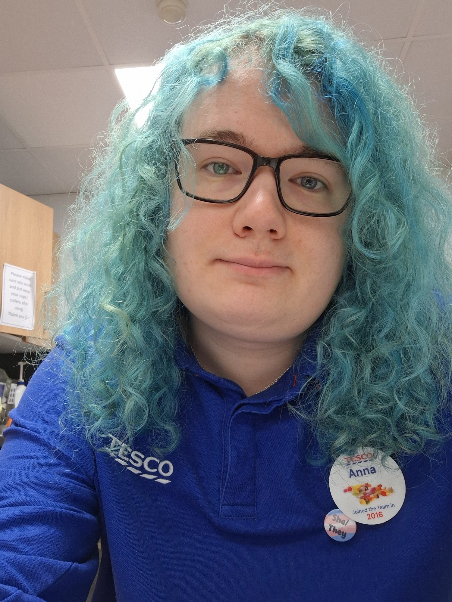 Anna with turquoise curly hair and in tesco uniform with a she/they badge