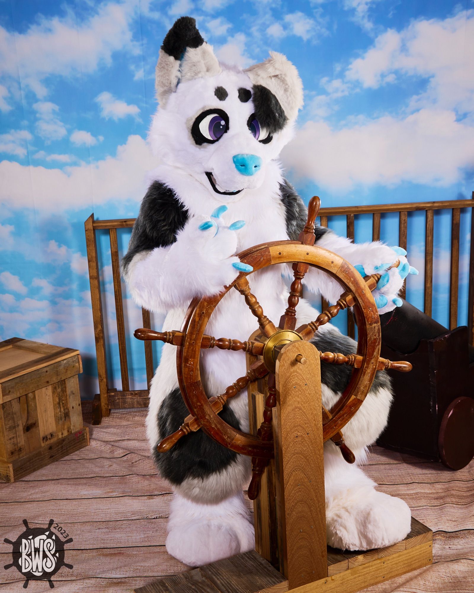 A white and gray dog fursuiter steering the ship’s wheel against a pirate-themed photo backdrop.