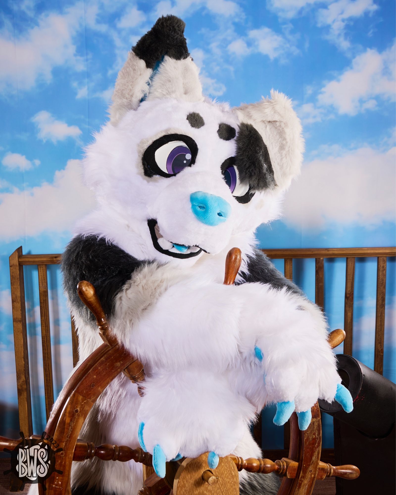 A white and gray dog fursuiter leaning on a pirate ship’s wheel.