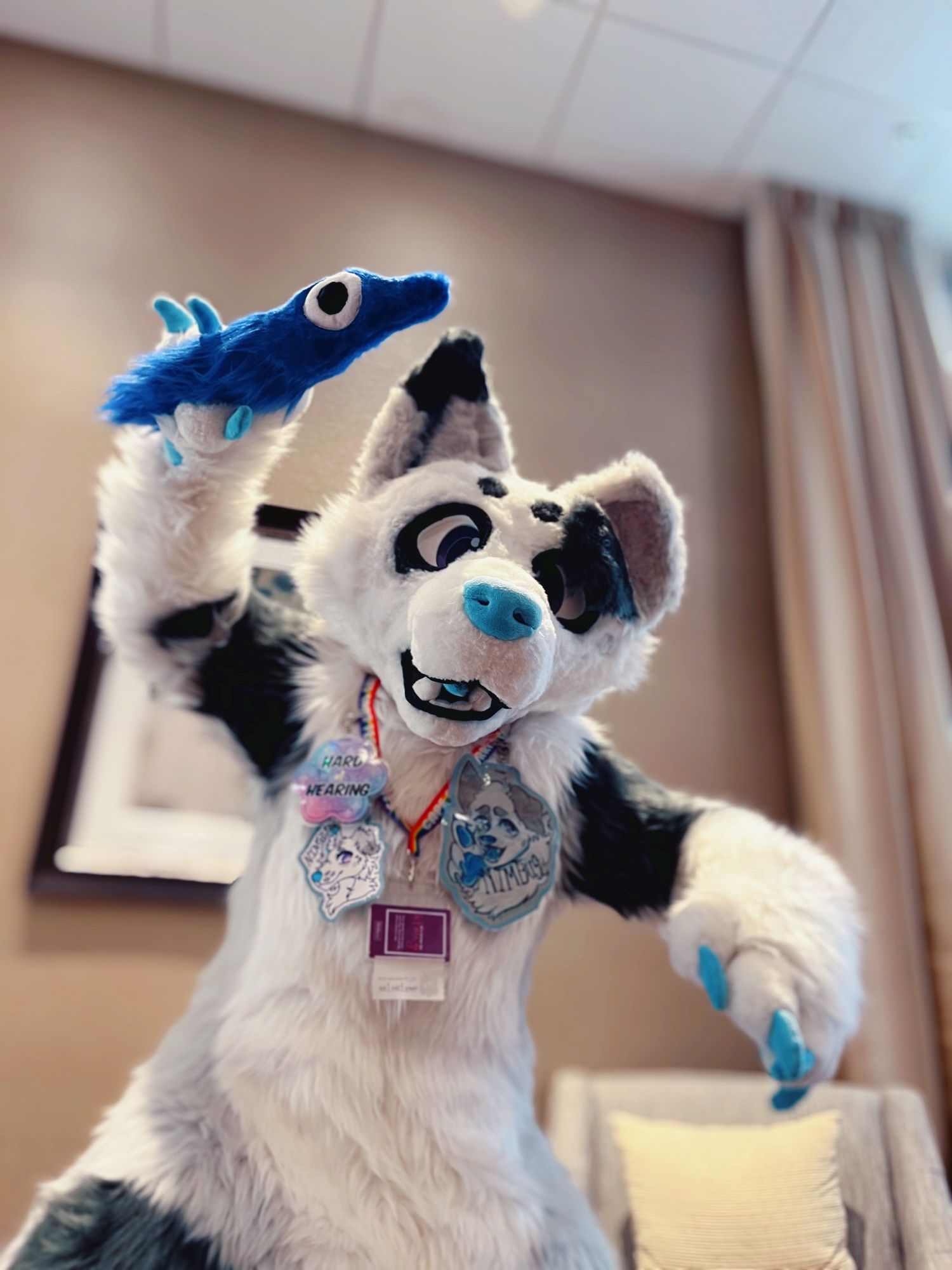 A white and gray dog fursuiter brandishing a fuzzy blue worm plush as if they are about to strike the viewer with it.