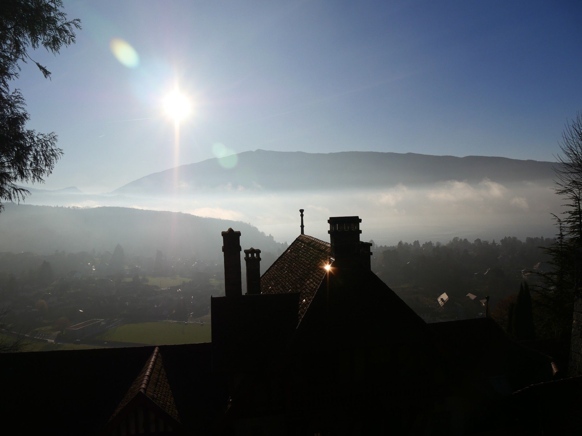 Le soleil qui fait un flare dans l'objectif, un reflet sur un toit du château au premier plan, le lac sous la brume