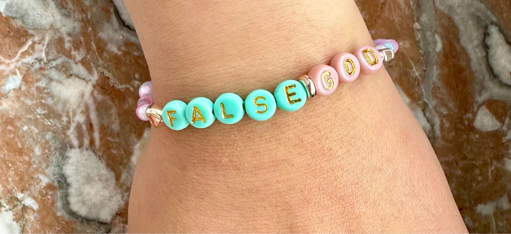 Nahaufnahme eines Friendship Bracelet an einem Handgelenk. Die Buchstaben bilden die Worte FALSE in hellblauen Perlen mit goldener Schrift und GOD in rosa Perlen mit goldener Schrift.