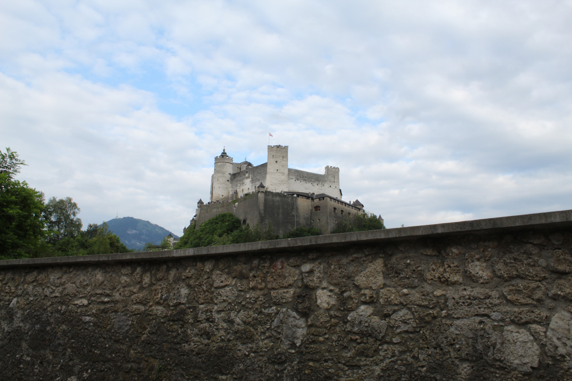 Josefsturm in Salzburg, Austria. The sky is mostly cloudy. A stony wall appears in the foreground with the hill and castle behind it.