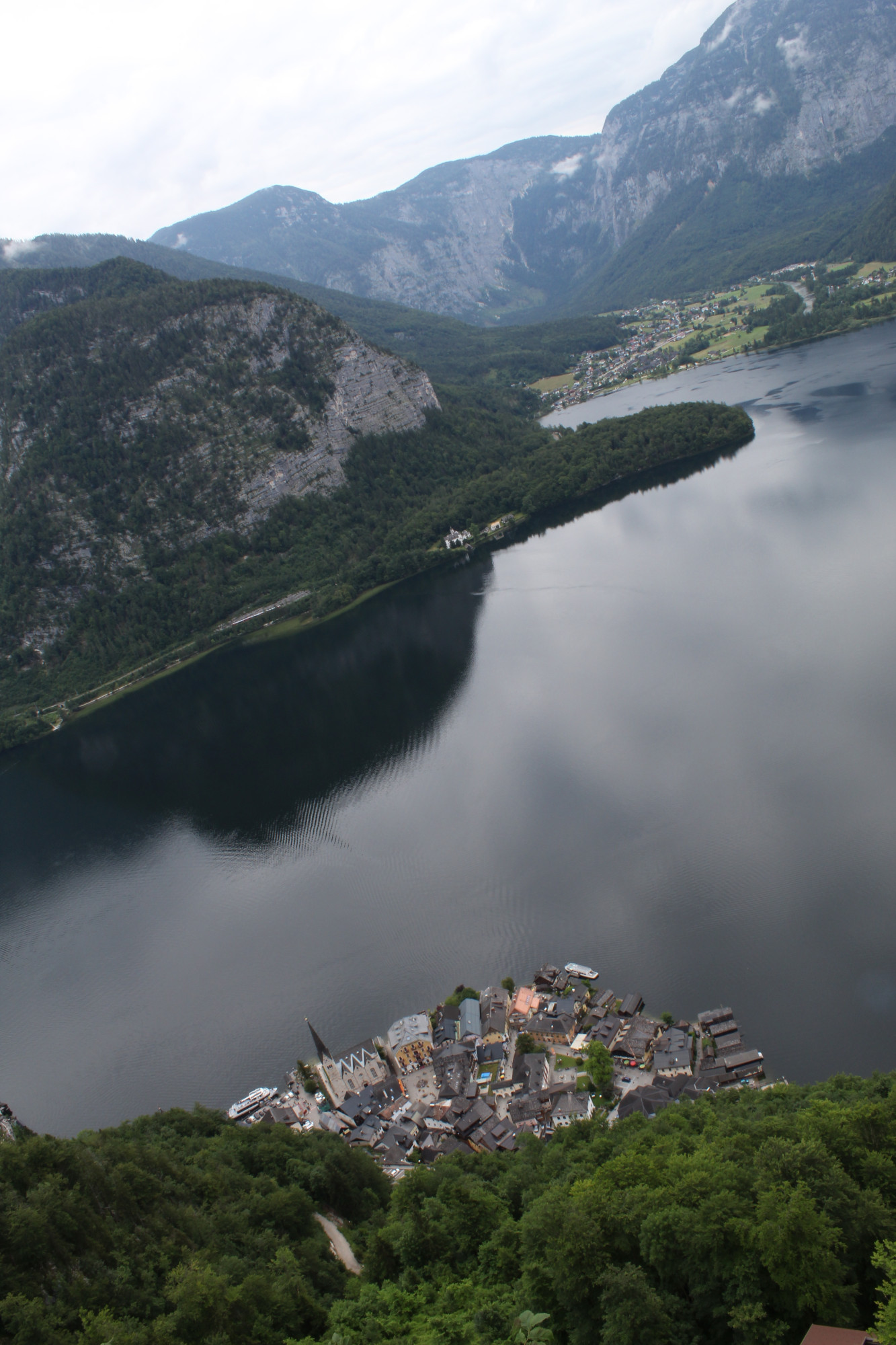 Hallstatt, Austria, taken from above. The viewing angle is skewed to include the lake and a valley in the distance. The reflection of the mountain across the lake is visible on the lake.