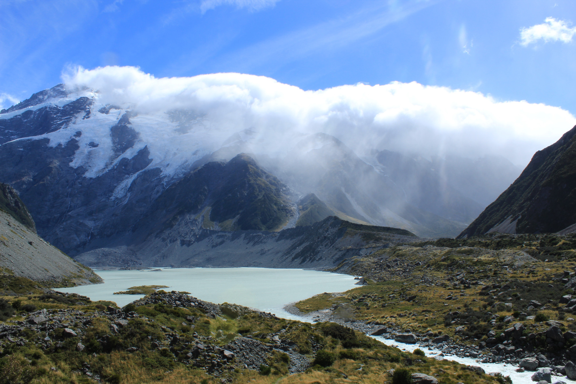 The Hooker Valley Track in Aoraki Mount Cook National Park, New Zealand. A tarn (glacial lake) with light blue water is visible in the foreground, with a rocky river leading out of it. The grass is a yellowish-green shade, and medium-sized rocks are strewn about. A translucent cloud obscures the valley in the right part of the background, but not the snowy mountain slopes to the left. The sky is clear save for a few wispy cirrus clouds.