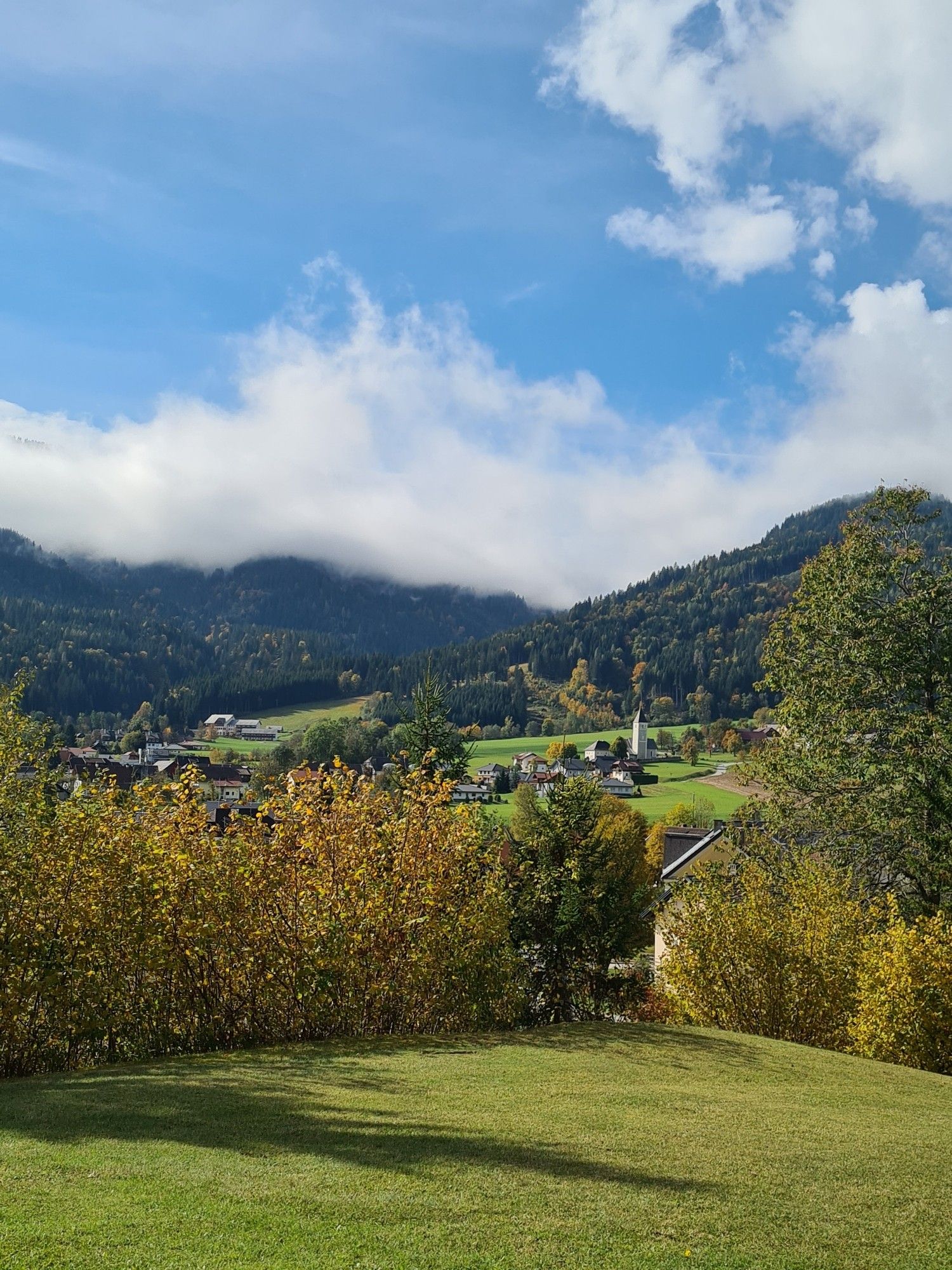 Blick in eine Herbstlandschaft, man sieht den Kirchturm einer Kirche, überall Herbstbäume. Überm Berg noch etwas hochhebel, aber alles in goldenes Licht getaucht