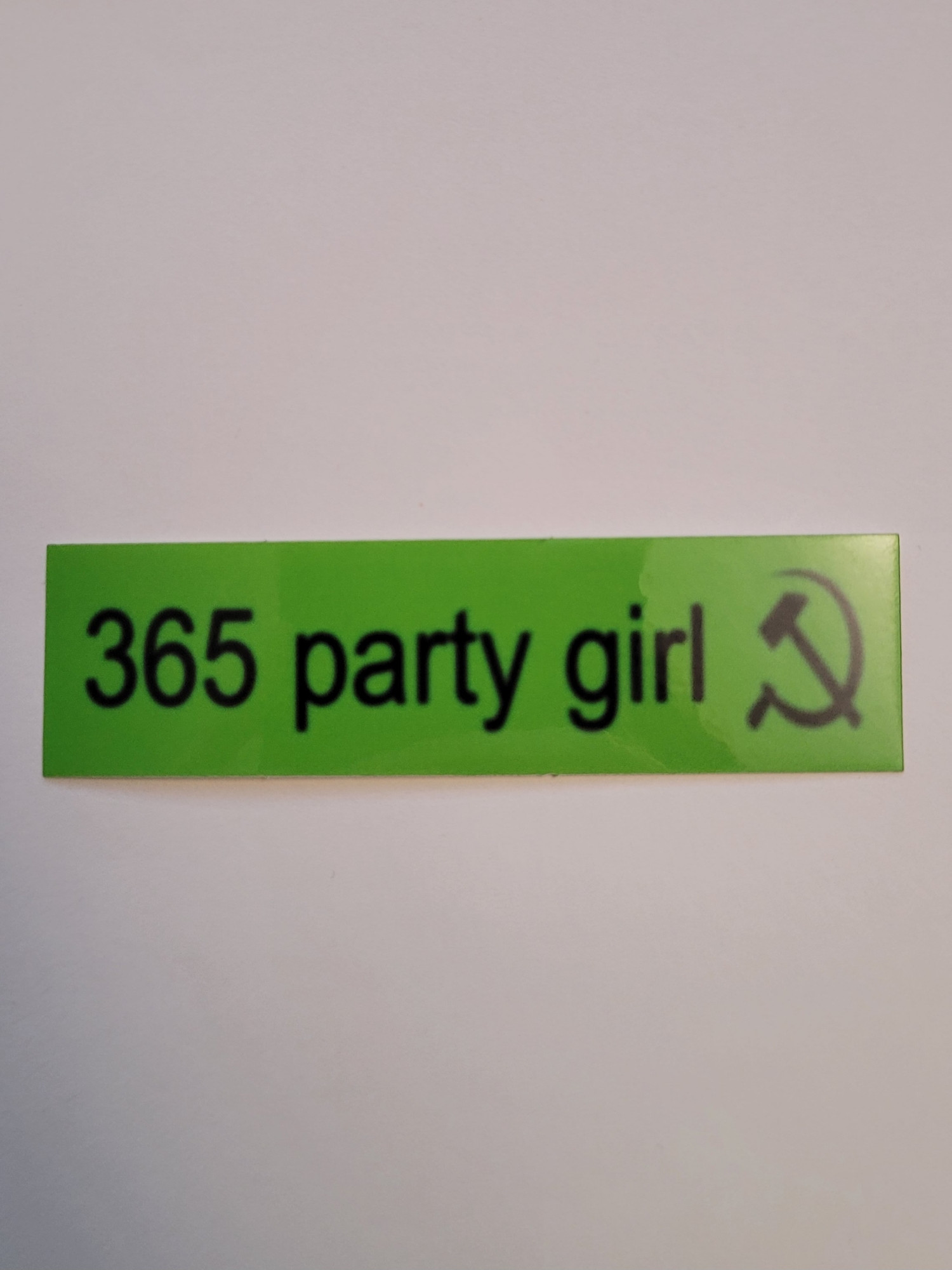 A neon green rectangular sticker with the black text "365 party girl" and a hammer and sickle. The text and hammer and sickle are blurry and slightly stretched to make everything seem a little taller and thinner.