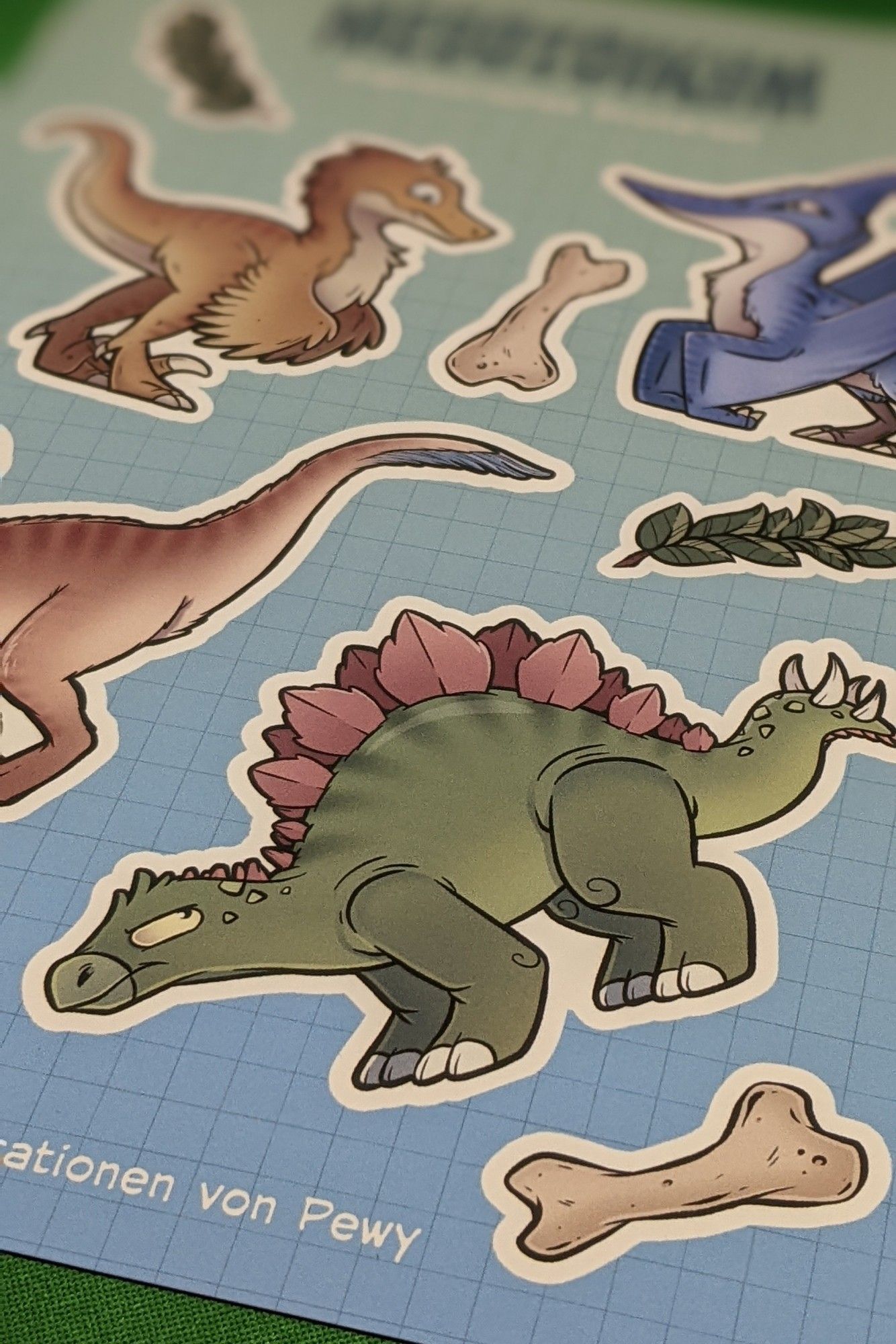 Stickersheet mit Comic Dinosauriern auf blauem karierten Grund. In den Lücken zwischen dej Dinos sind Knochen und Äste eingezeichnet.