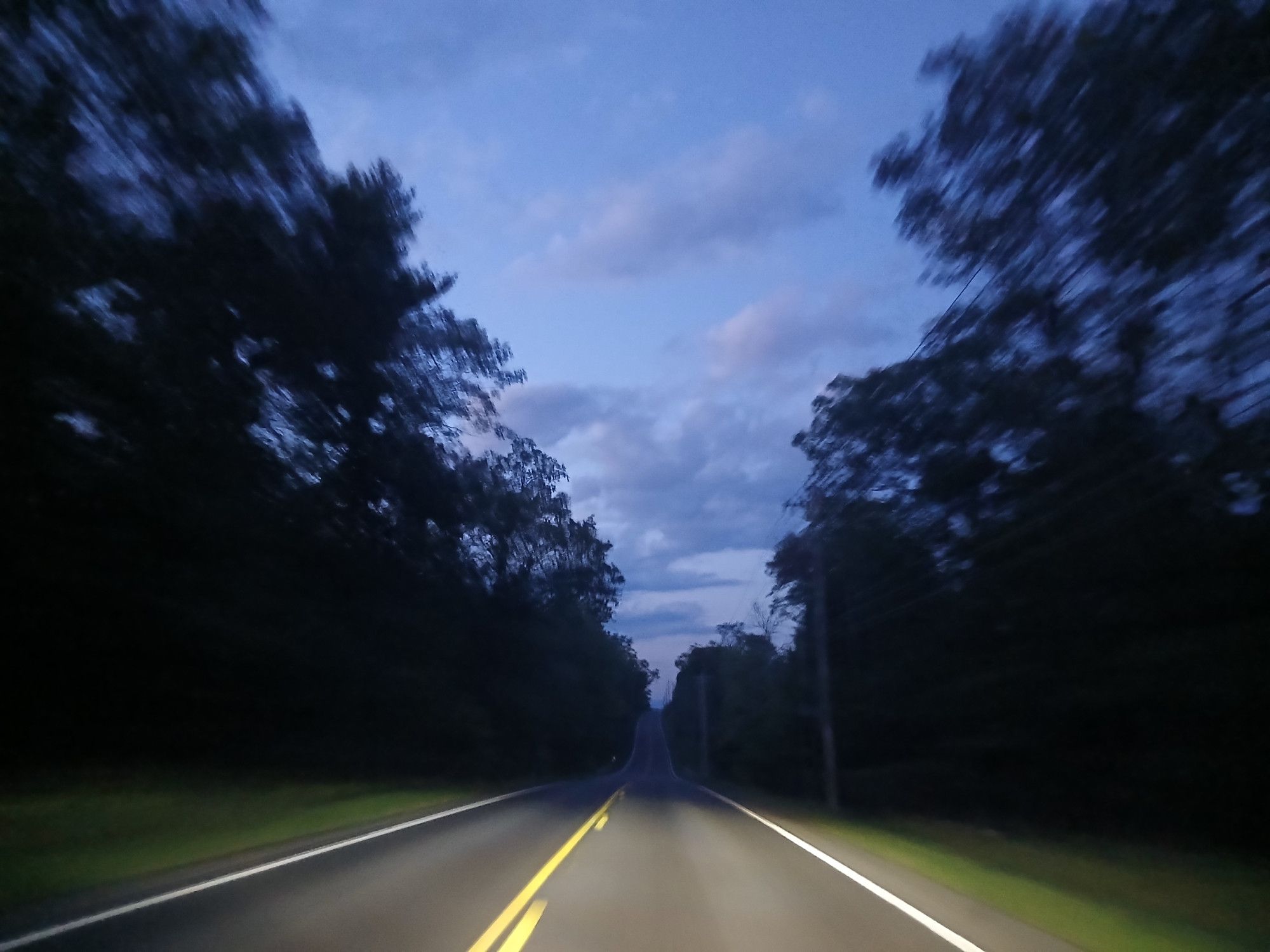 Country road with forest on both sides of the road. Sky starting to get darker as nighttime gets closer. Headlights of the car light the road ahead (white and yellow painted lines on the road).