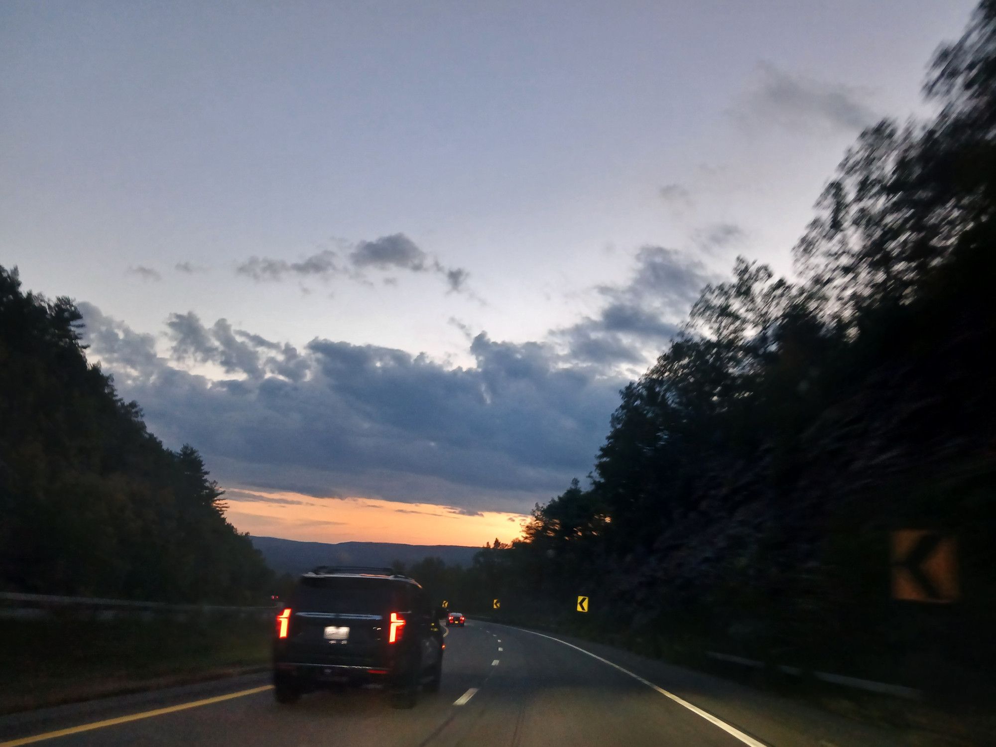 Driving over a mountain ridge with a valley below. Mountain rocks on both sides of the road. Setting sun creates a golden hue above some mountains in the distance, some clouds over the golden hue and then clear blue sky.