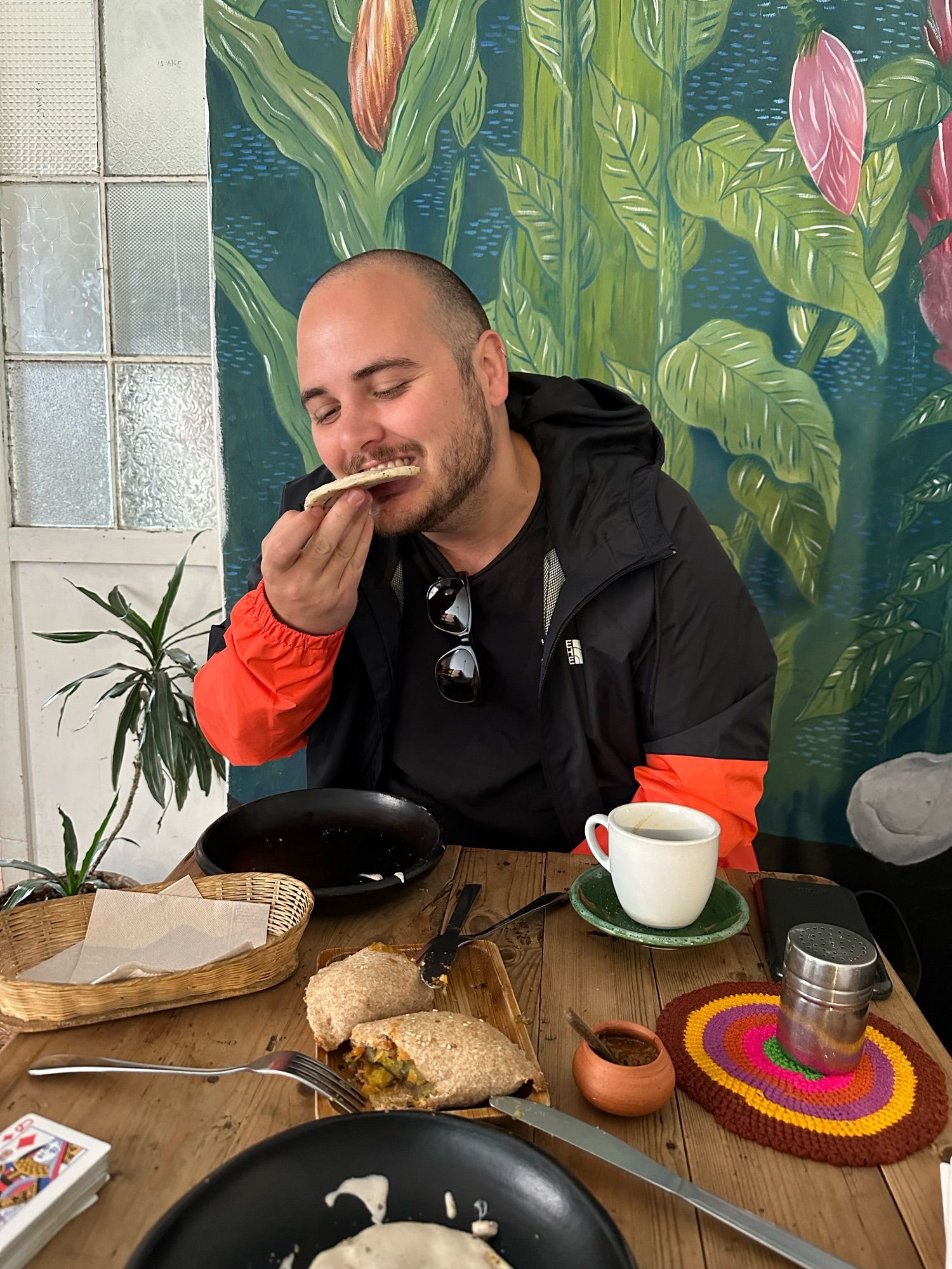 Me eating an arepa covered in a kind of cheese sauce. There’s a couple of big empanadas half eaten on the table in front of me, plus a coffee and playing cards. A houseplant is behind me and a nice mural is painted on the wall behind me - flowers and leaves.