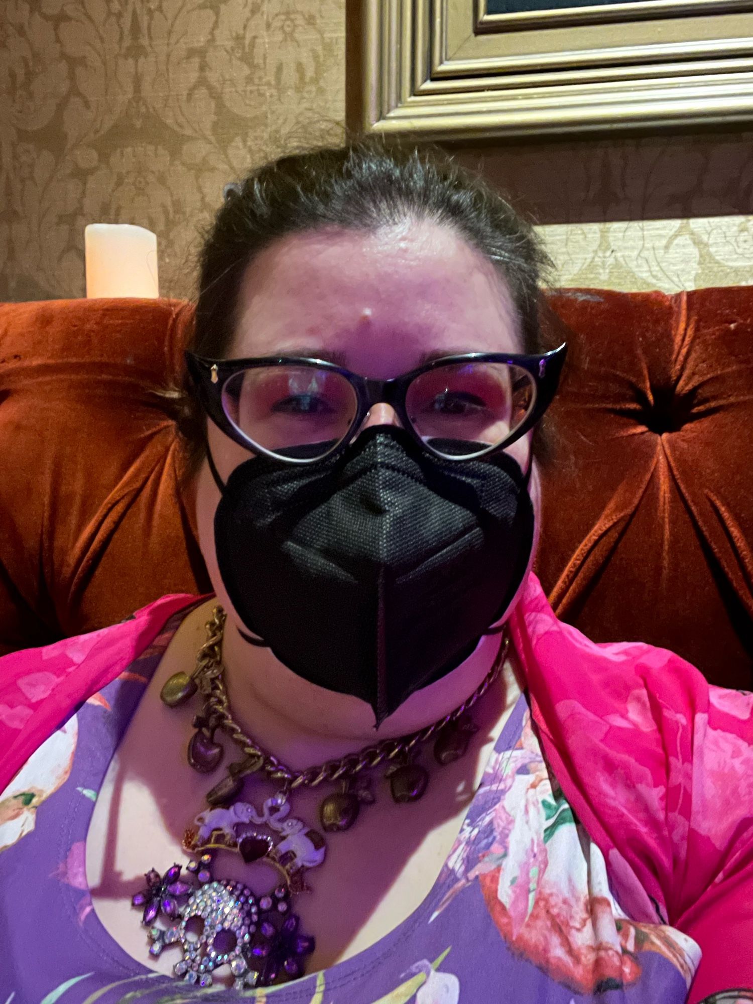 Selfie. Fat white brown haired person wearing a bright pink cardigan and a tropical skull print dress in purple and pink, with a big glitter skull necklace. I am wearing cateye glasses and a black n95 mask.