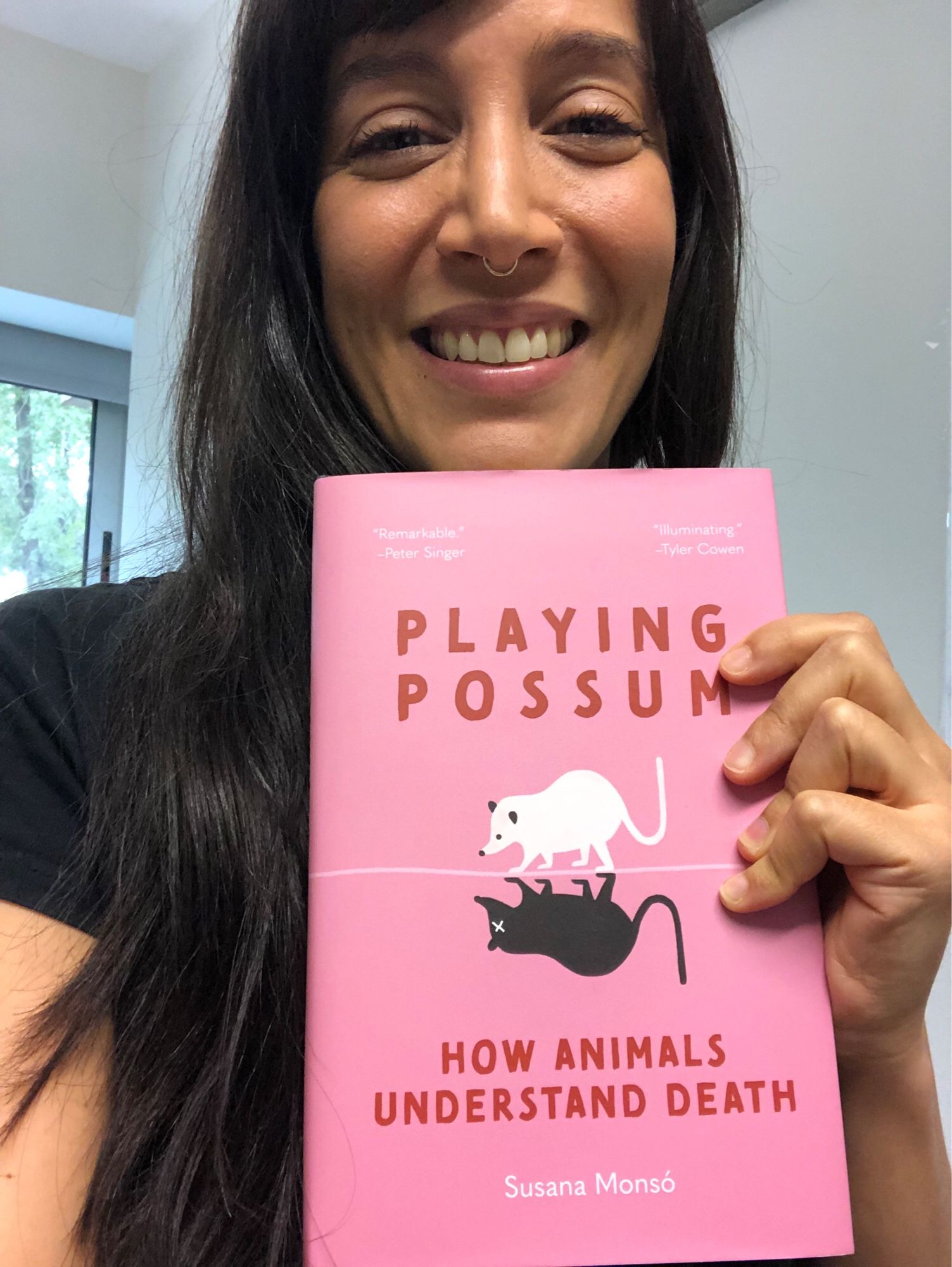 A picture of me looking ecstatic holding a copy of Playing Possum: How Animals Understand Death. The cover is bubblegum pink and features a white opossum with an upside-down dead counterpart.