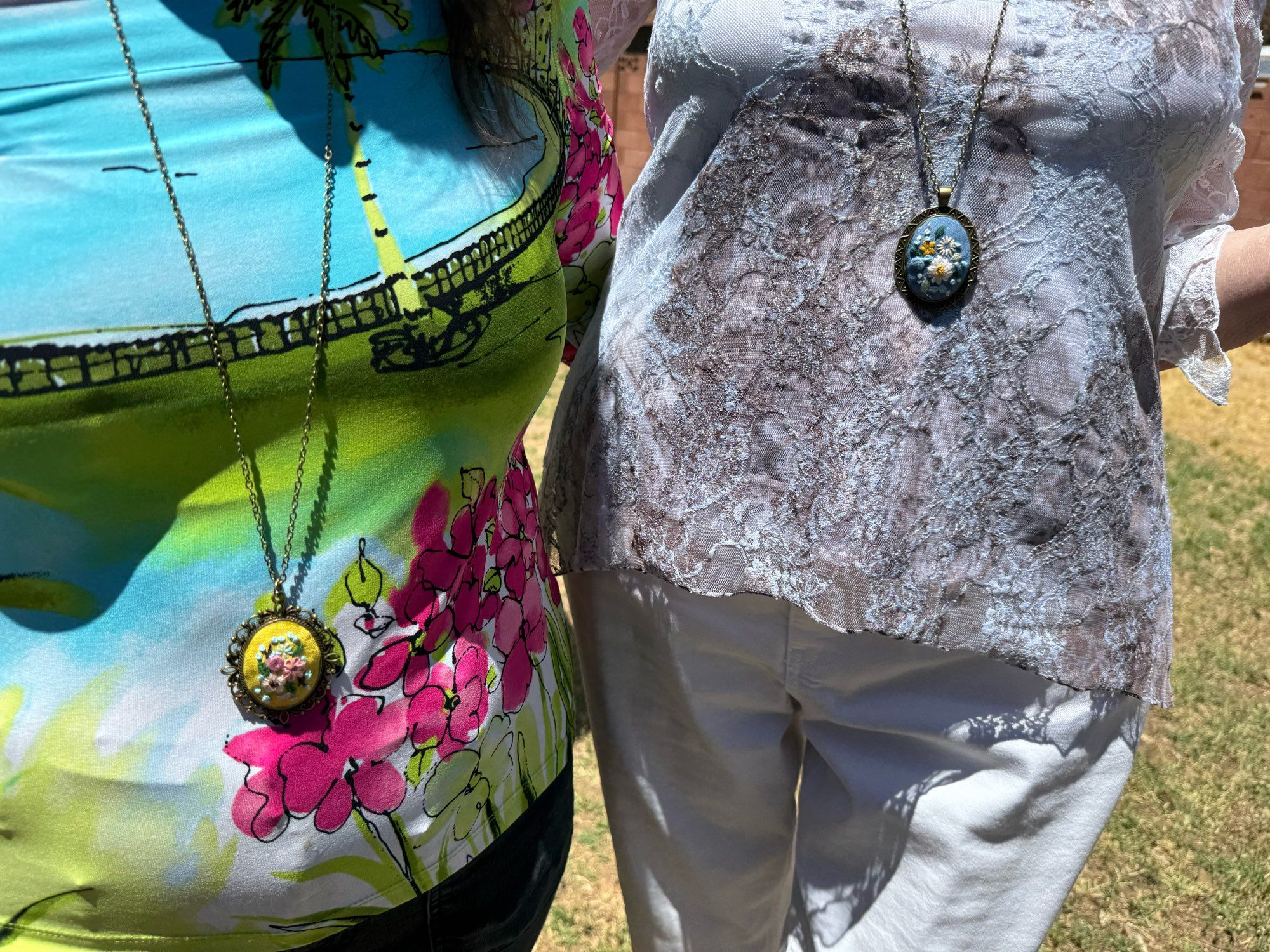 Torsos of two women, one wearing white slacks and a gray shirt, the other wearing black pants and a colorful shit. They have metal pendants around their necks with tiny flower embroideries.