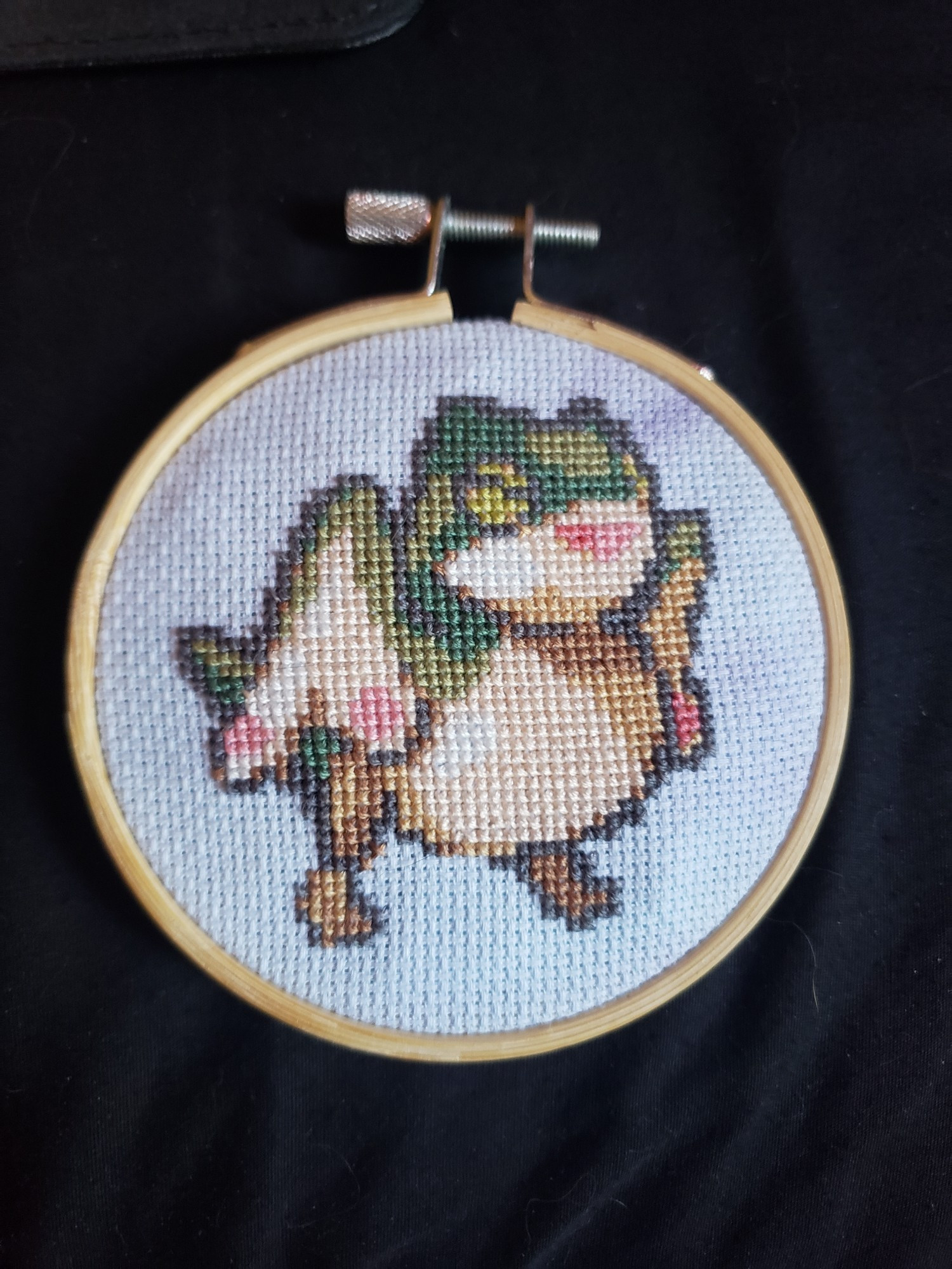 A cross stitched baby pukei pukei based on pixel art from gandalfsoda. It is displayed in a 3.5 inch embroidery hoop. 