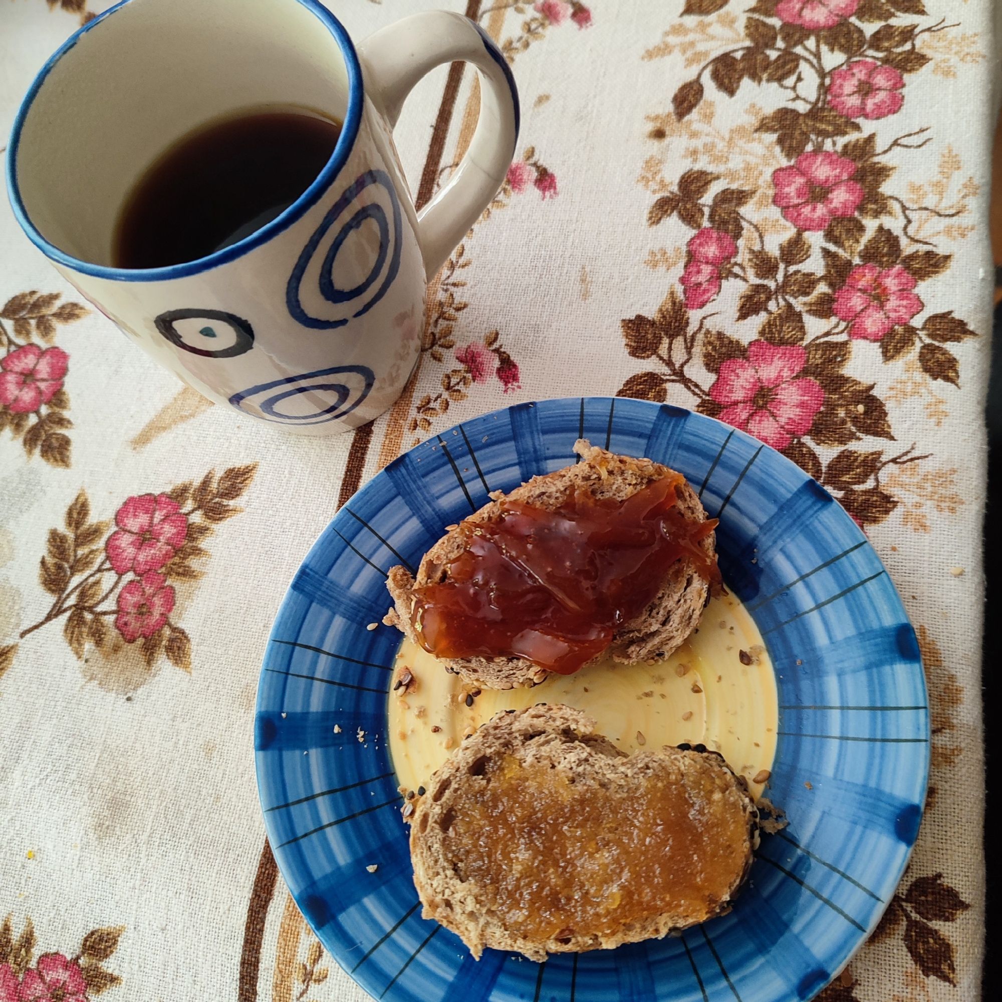 Xícara com café preto e pratinho com duas fatias de pão de grãos, uma com mel e outra com geleia de laranja, sobre toalha florida.