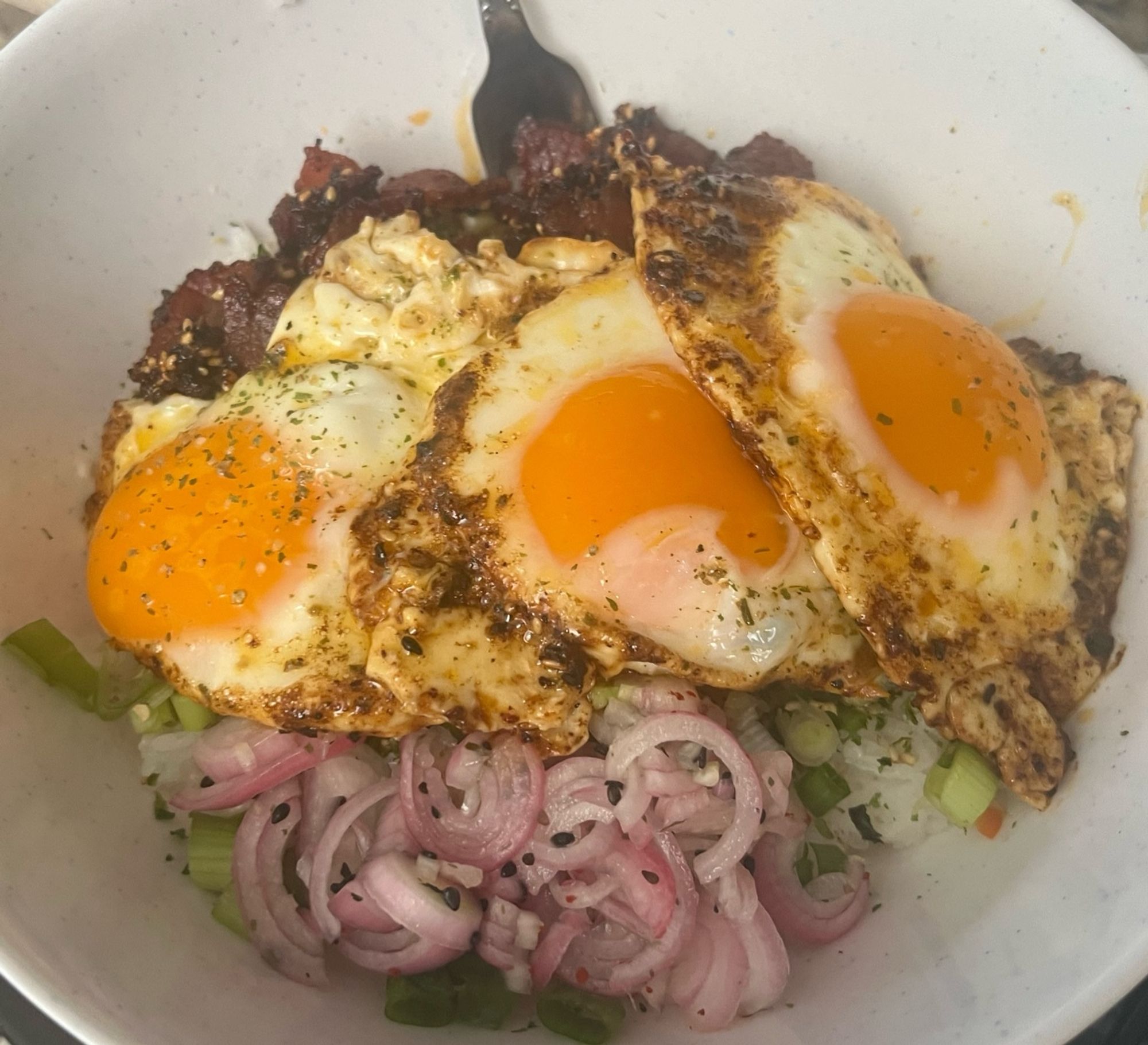 A bowl of garlic rice with furikake, with sweet sesame bacon, quick pickled shallot, green onion, Thai chili, and 3x fried eggs