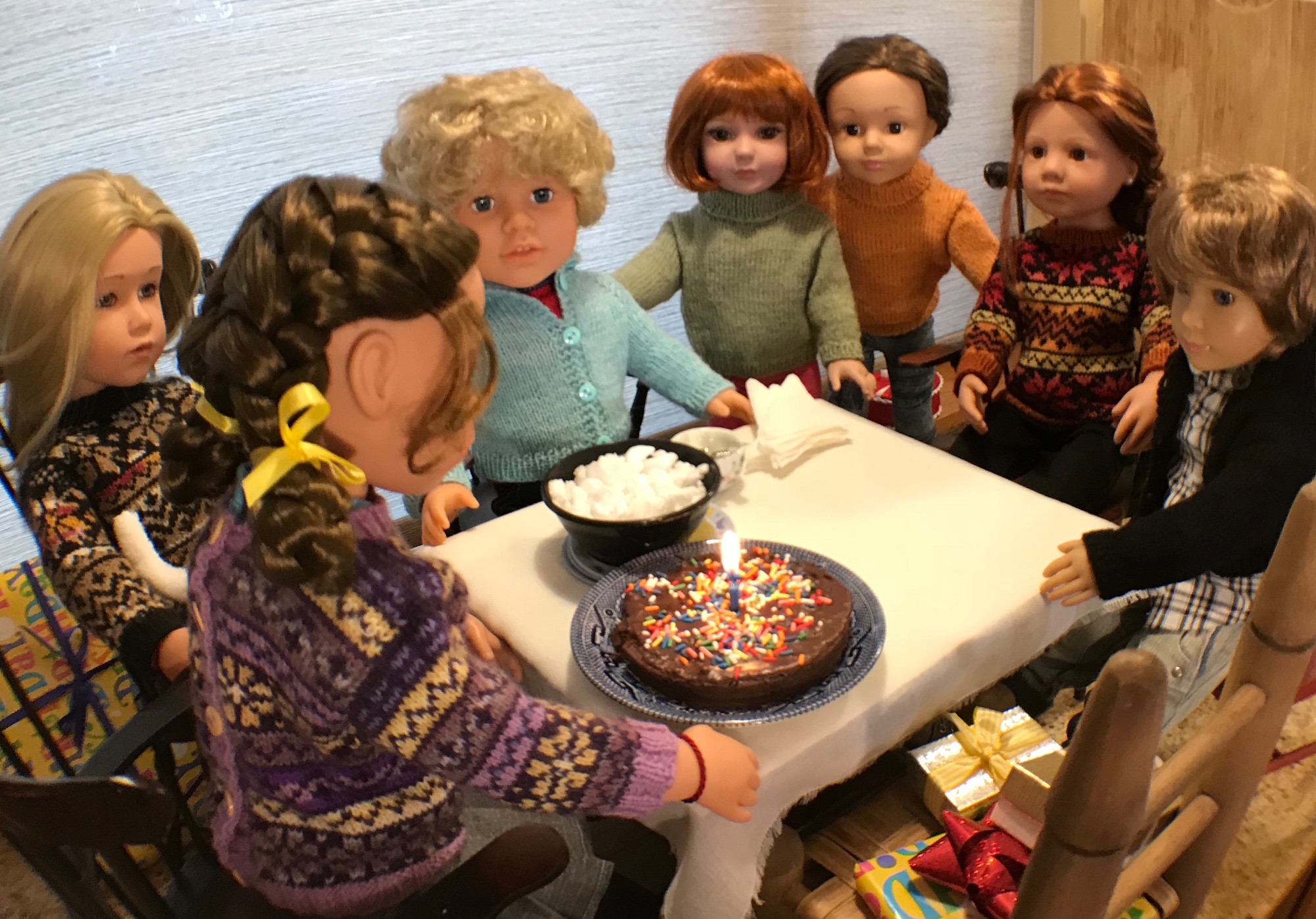 Dolls are seated or standing around a table. On the table is a small cake with one candle and a bowl of what appears to be popcorn. One chair is filled with wrapped packages.