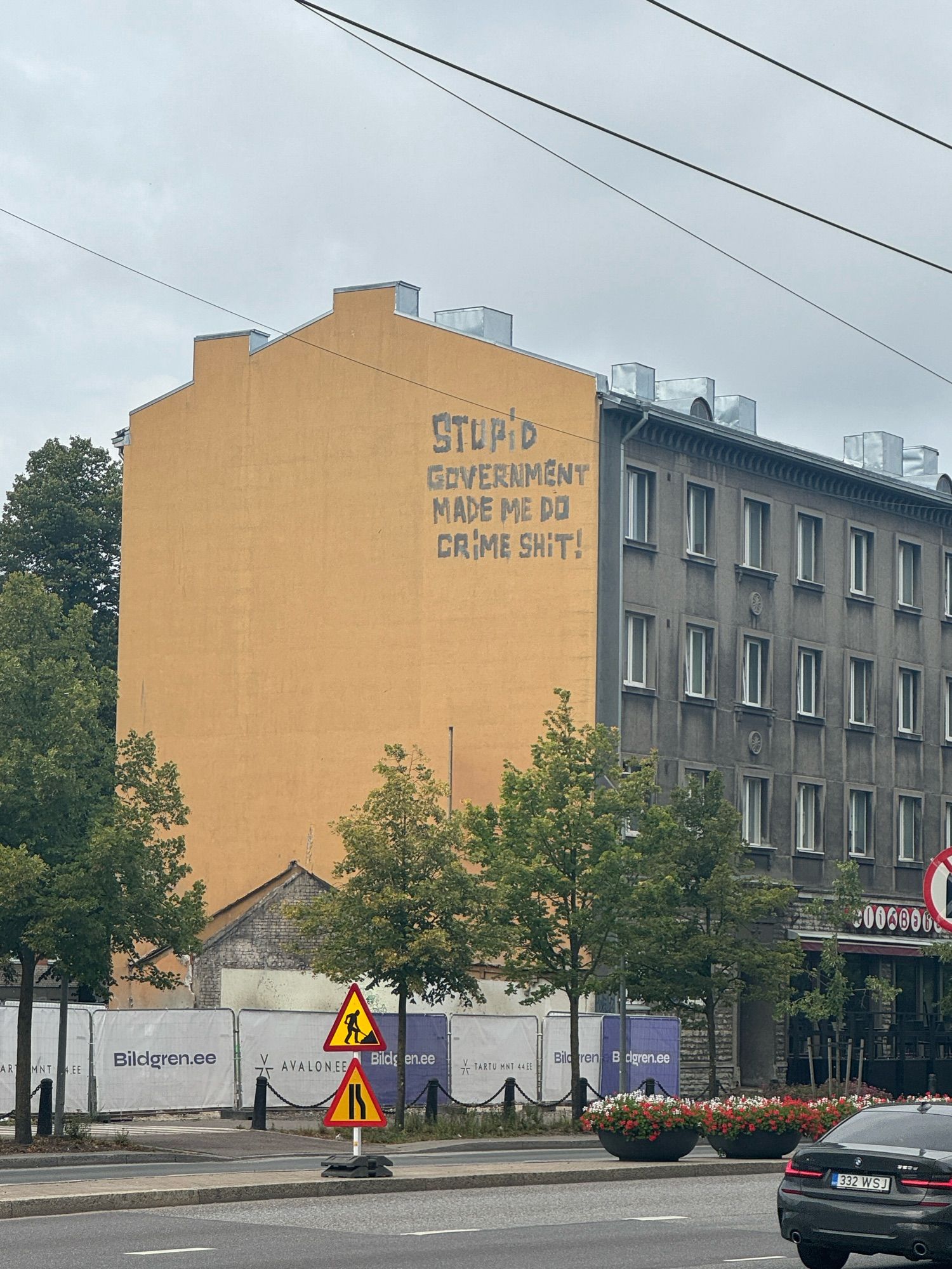 A four-story building in Tallinn, featuring a blank yellow wall on one side. Way up at the fourth floor, someone has painted “STUPID GOVERNMENT MADE ME DO CRIME SHIT!” in gray. It slaps, ngl