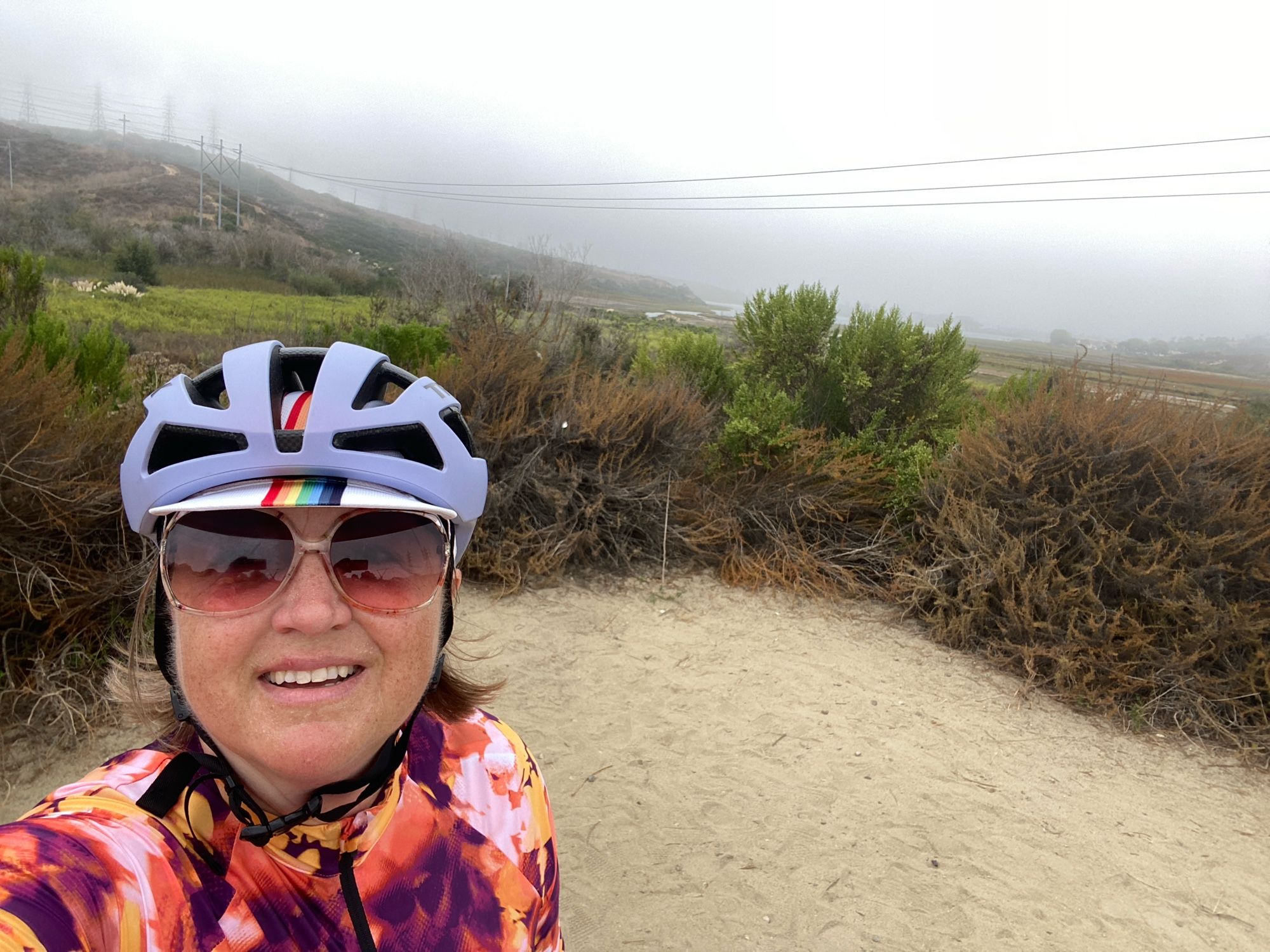 A selfie of me in bike helmet and sunglasses, standing on a dirt trail, looking west over the Agua Hedonia lagoon. There’s so much mist you can’t see the ocean.