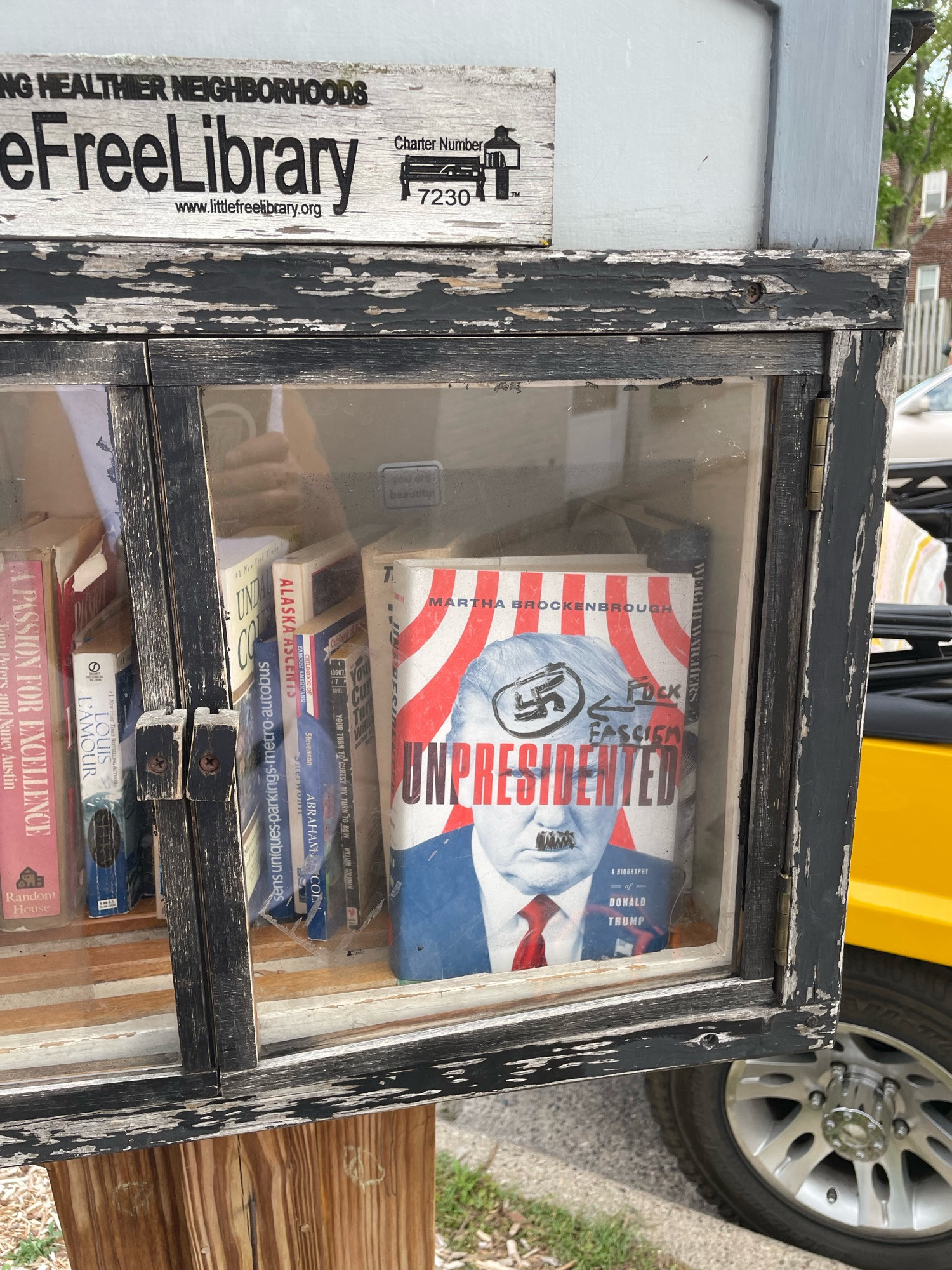 Wooden framed glass door of little free library. A book facing out is “Unpresidented”— picture on cover is of Trump and someone has put a Hitler mustache and swastika on him and written “fuck fascism” in black marker