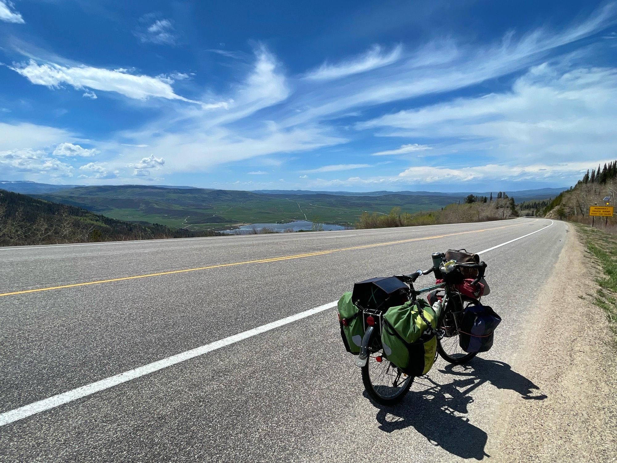 Photograph of a heavily loaded Surly Long Haul Trucker bicycle with full front and rear panniers, a solar panel over the gear on the rear rack, and gear strapped to the handlebars over the front rack, parked along the side of a road heading down a mountain to a green valley with a lake near Steamboat Springs, Colorado. There are white clouds stretching across a blue sky.