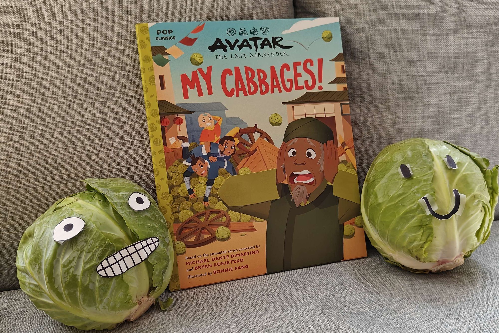 The cabbage merchant picture book My Cabbages! beside one cabbage with a smiley face taped on it and one cabbage with a cursed grimace face taped on it.