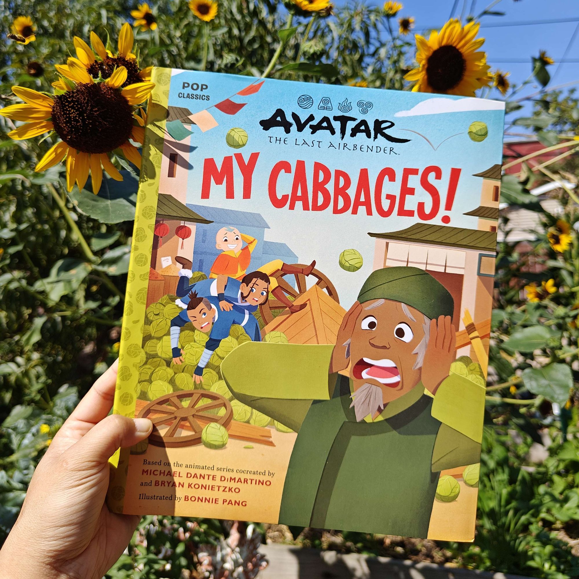 Picture book titled MY CABBAGES with the cabbage merchant clutching his head in distress as his cabbages tumble out of a cart. The book is held up in the sunlight against sunflowers