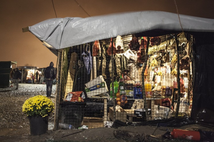 Photographic image of a grocery store in a tent in the Calais 'Jungle' in 2015, with a bucket of yellow flowers to the left of the enclosed shop.