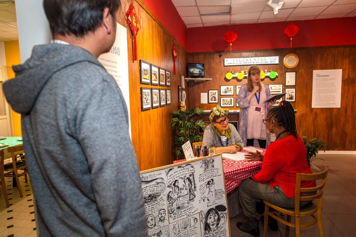 Interior of a Welsh Chinese take-away shop, the Lucky Star, with images of good and a table, two seated attendees discuss the display.