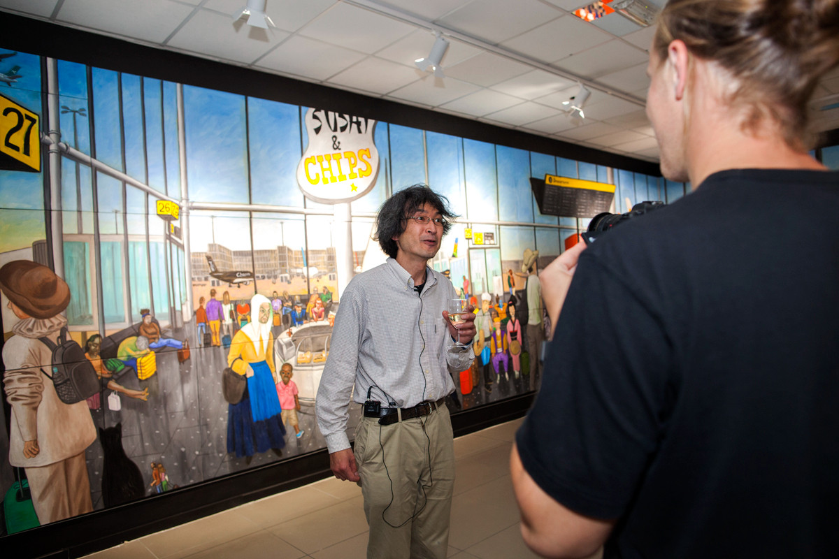 The artist Jiro Osuga stands in front of his colourful imagined airport departure lounge, at the launch of All Our Stories, engaging with the audience.