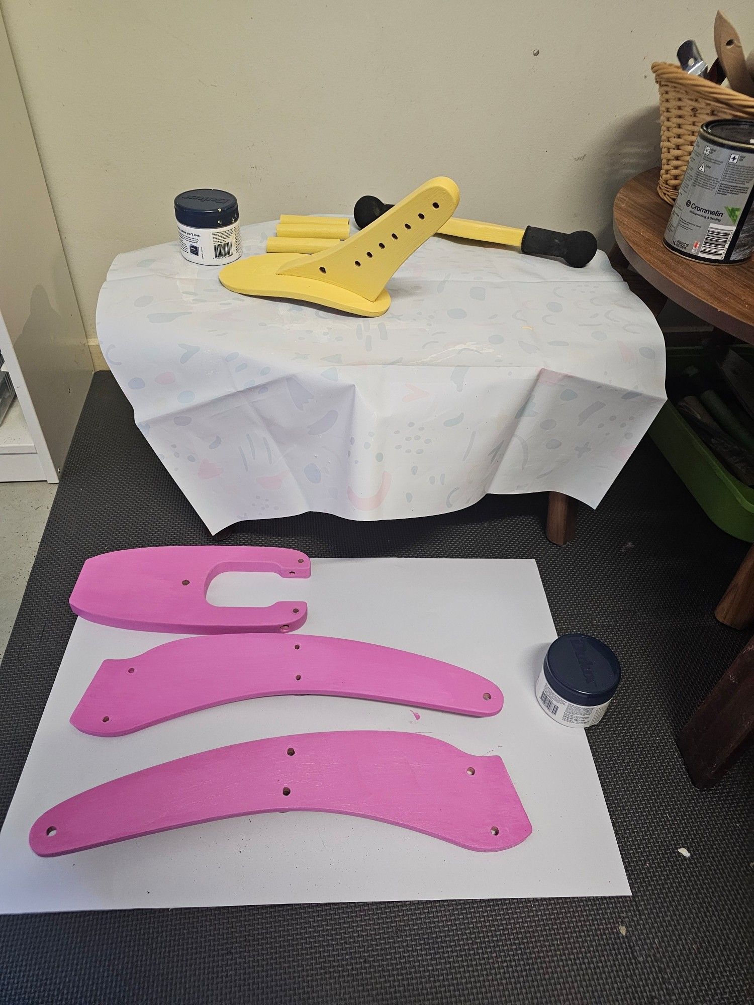 Image show a table with a white cover and dismantled parts of a bike painted bright yellow.

On the floor in front of the table is a white sheet of paper with dismantled bike parts painted in bright pink.