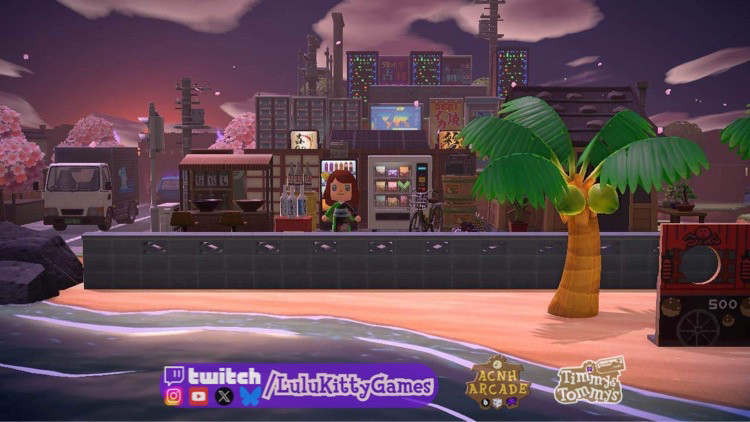 An animal crossing game virtual island build towards the end of sunset. In the foreground, a beach with a coconut tree. In the mid ground is a city street scene and pink cherry blossom trees. Simulated apartment buildings and utility poles rise in the background.