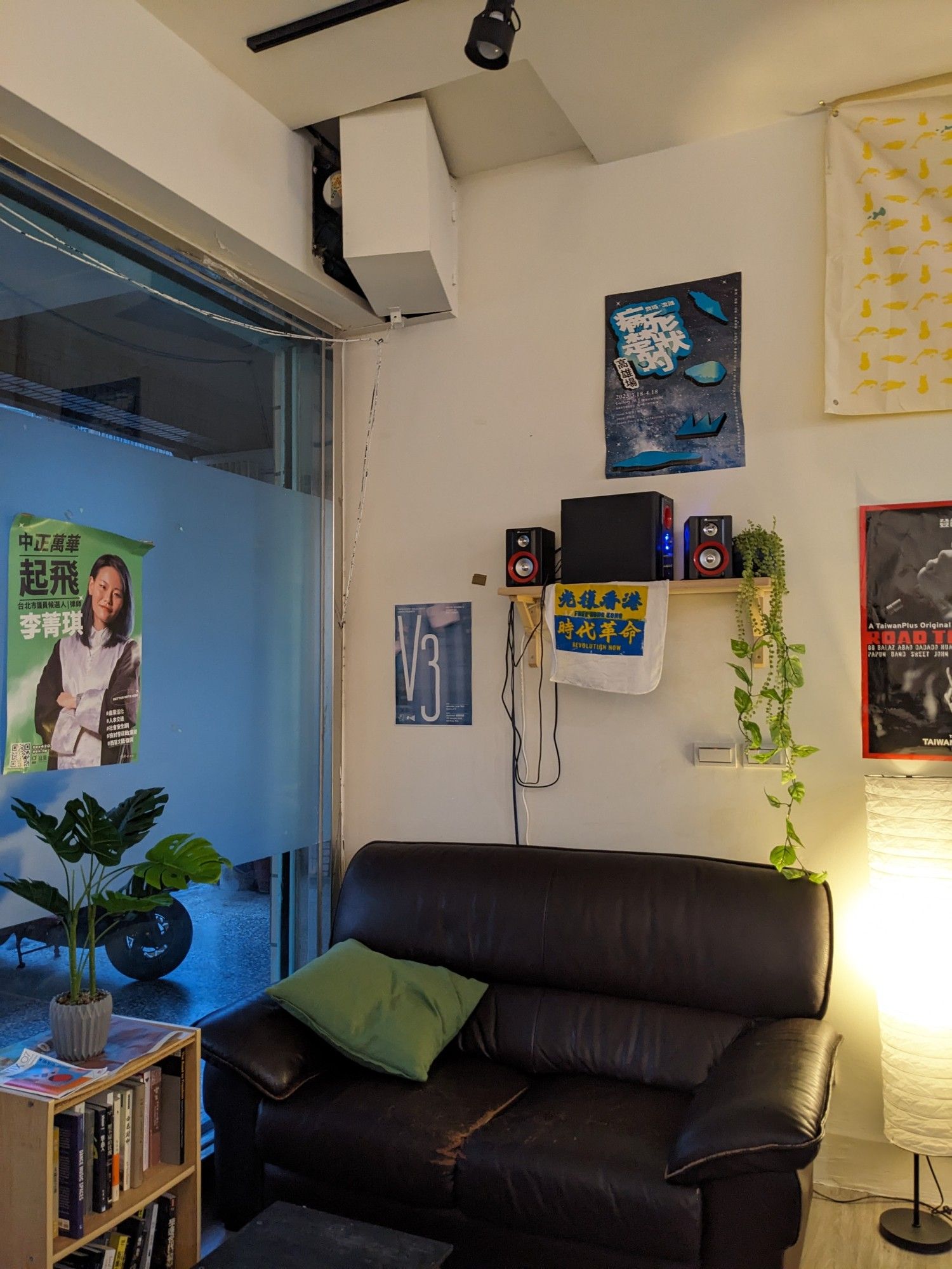 Cosy sofa area with plants streaming down from the stereo shelf. lighting is creamy and welcoming, pillow and plants are green. there is a hand dyed sign that says 光復香港 時代革命 in the colours of the Ukrainian flag