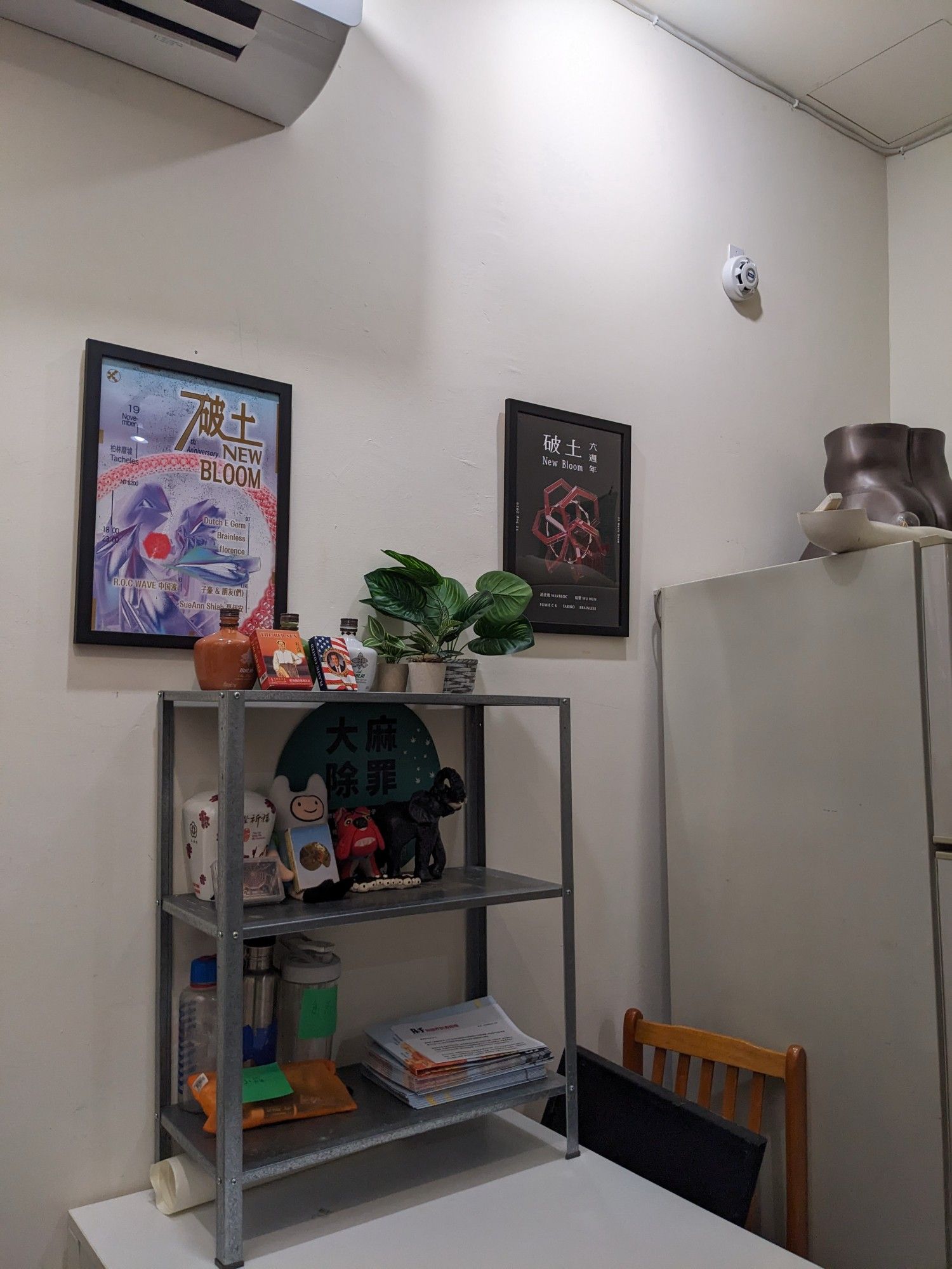 Steel cabinet added to white table, with various fake plants and knick knacks organised in them. New bloom anniversary posters decorate the wall behind the shelf 