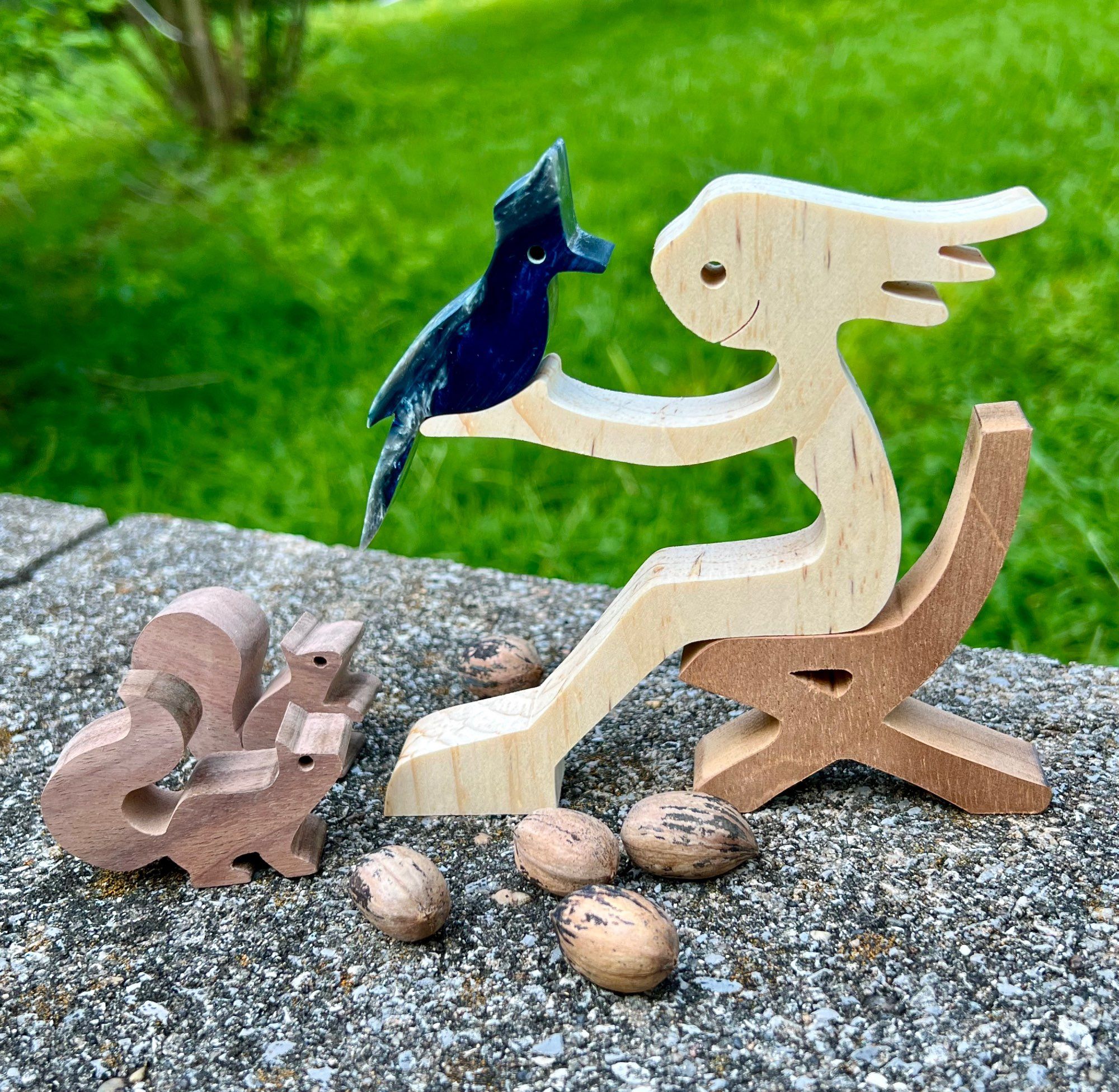 Wood stick figure of smiling female sitting in chair. A Steller's Jay sits in her outstretched hands while 2 squirrels watch intently. Tiny pecans surround them.