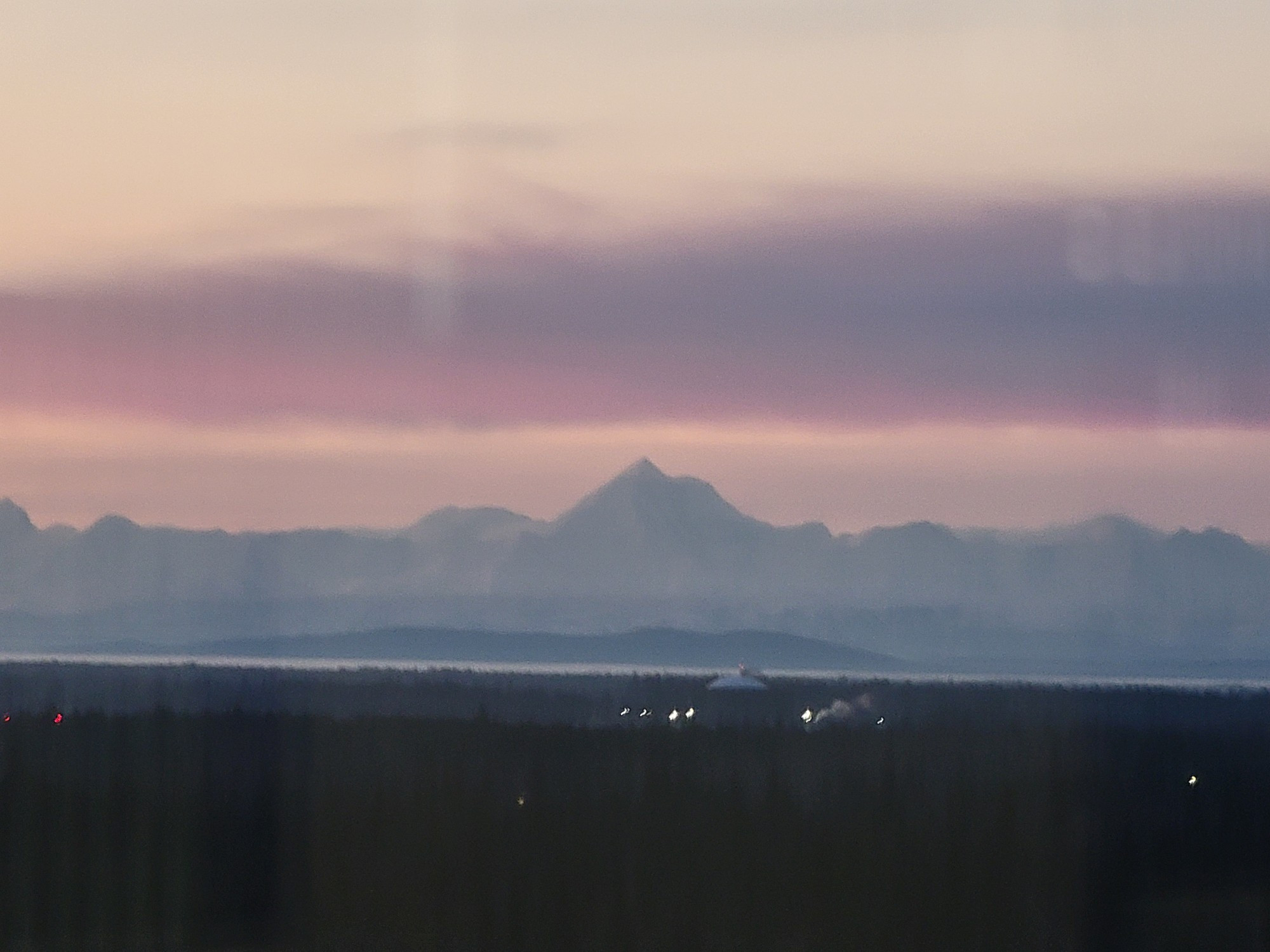 Hazy pink skies and low clouds over the mountains. A small building and lights can be seen amongst the dark forest in the foreground.