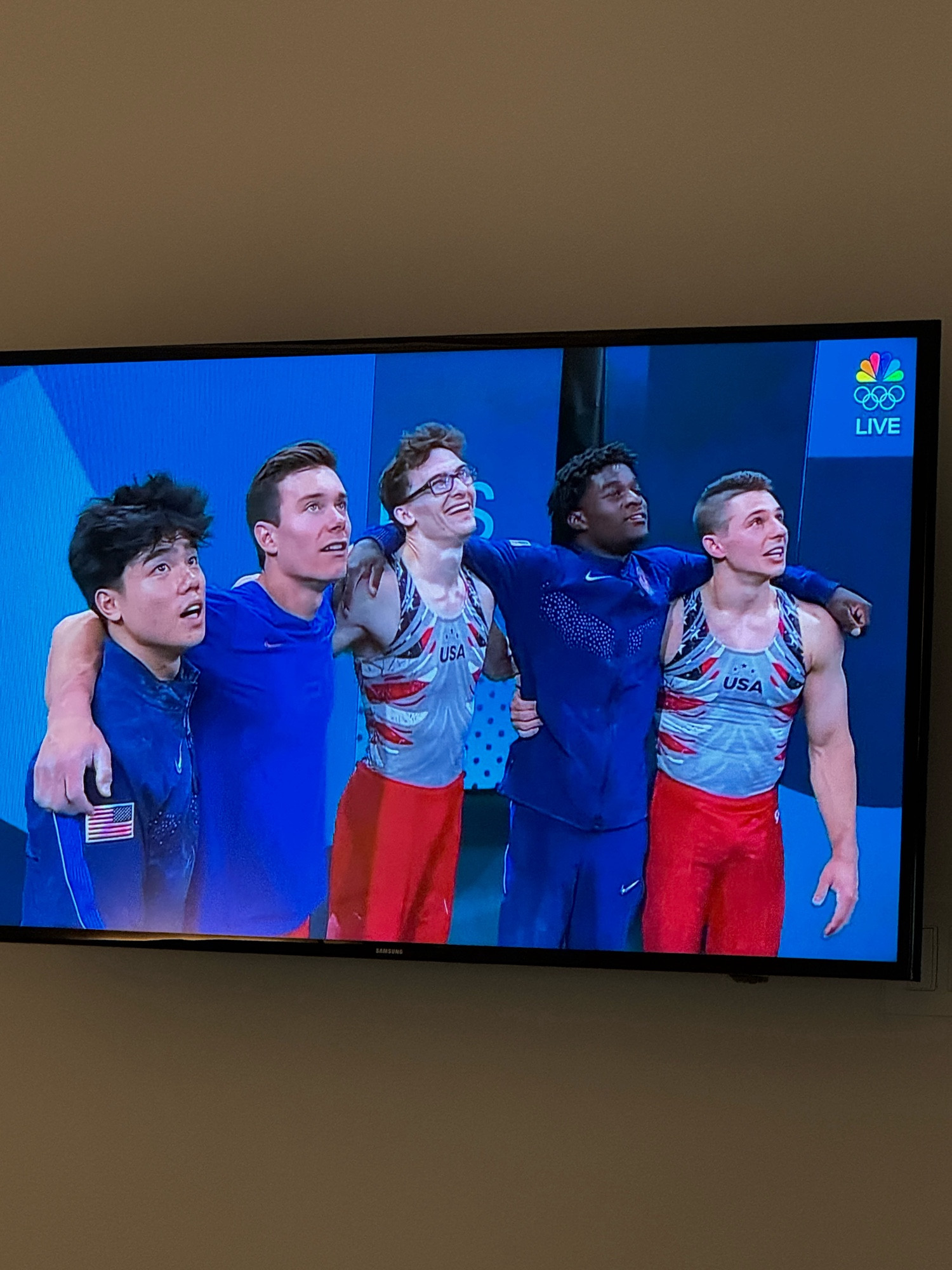 The USA men’s gymnastics team with their arms around each other’s shoulders while they await a score.