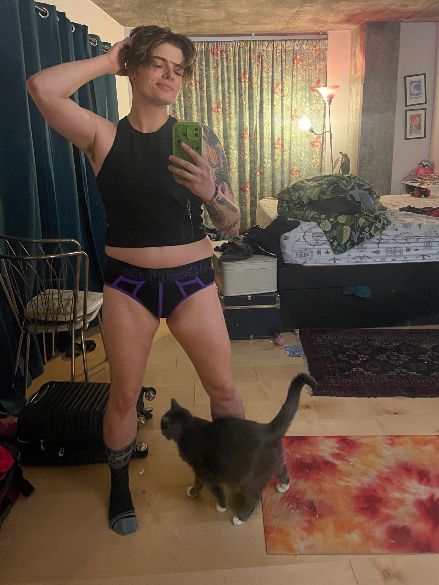 Micki wearing a black tank top and black and purple briefs. Coppie the cat is walking by in the frame